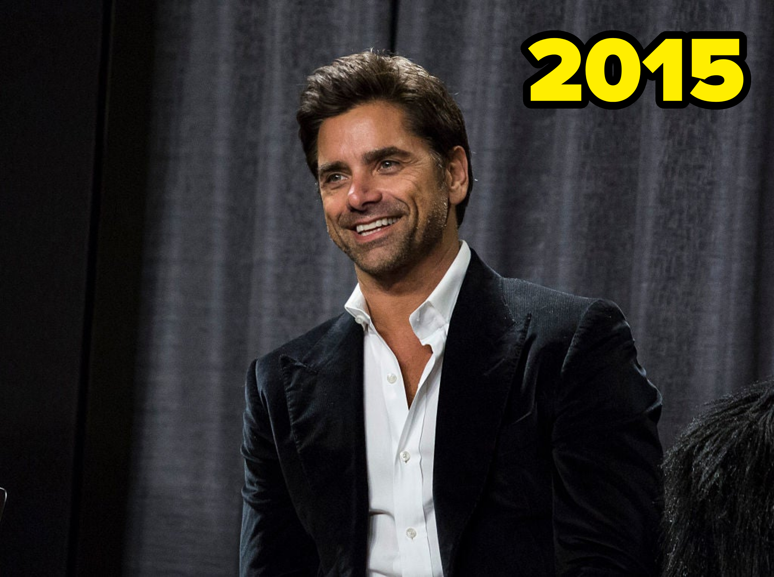 John Stamos is smiling while wearing a black suit jacket and white shirt in front of a dark curtain backdrop
