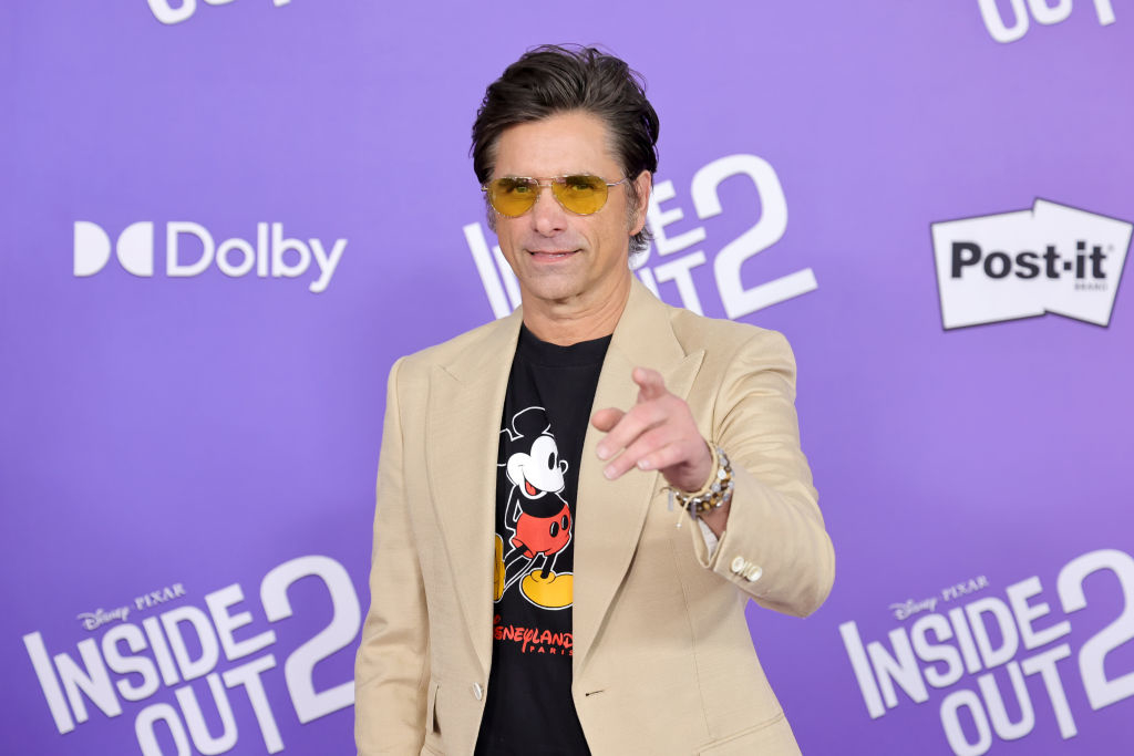 John Stamos at the premiere of Inside Out 2, wearing a beige blazer over a Mickey Mouse t-shirt, posing and pointing towards the camera