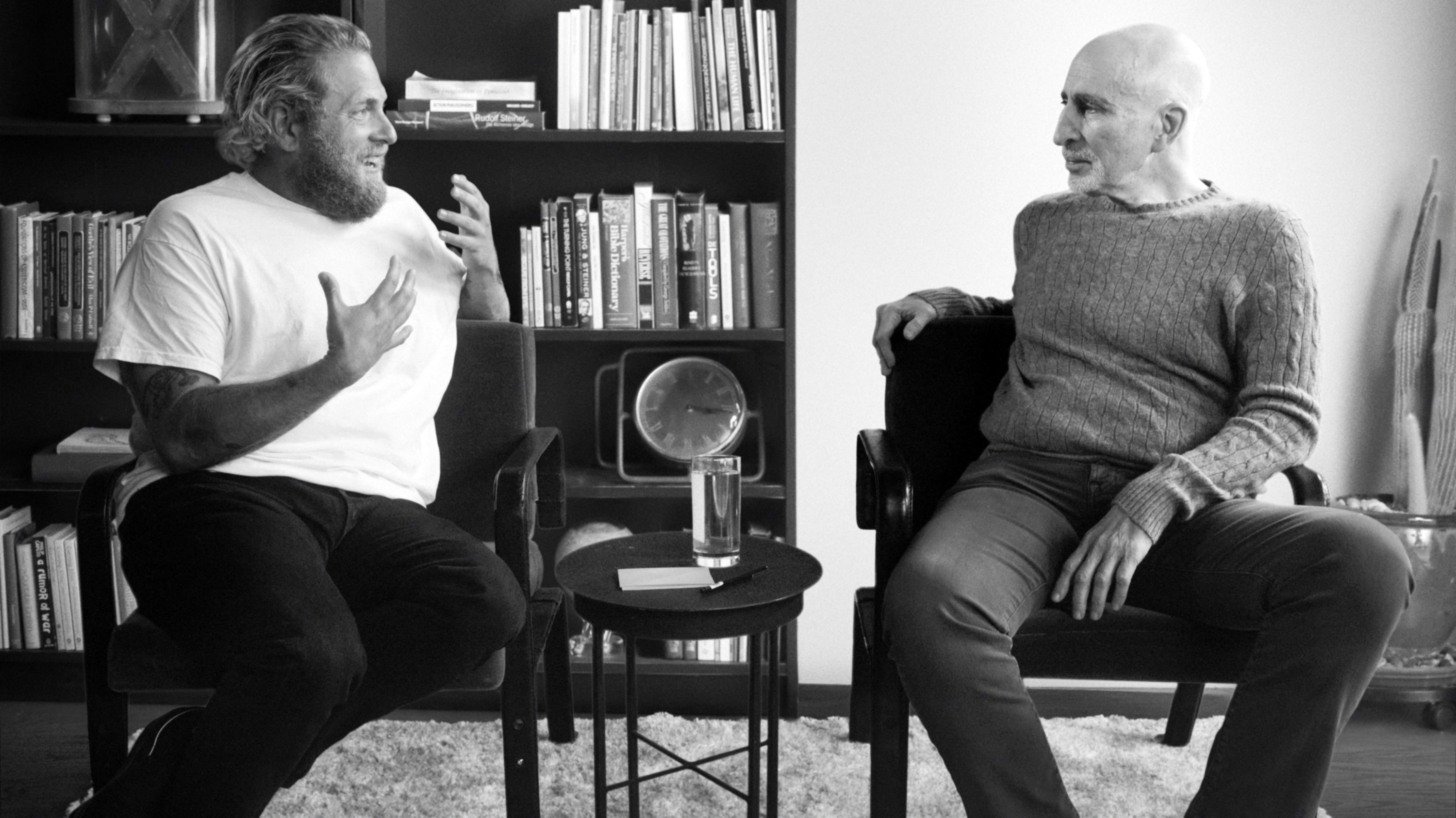 Jonah Hill and Phil Stutz sitting and conversing in a room with bookshelves in the background. They are engaged in an animated discussion