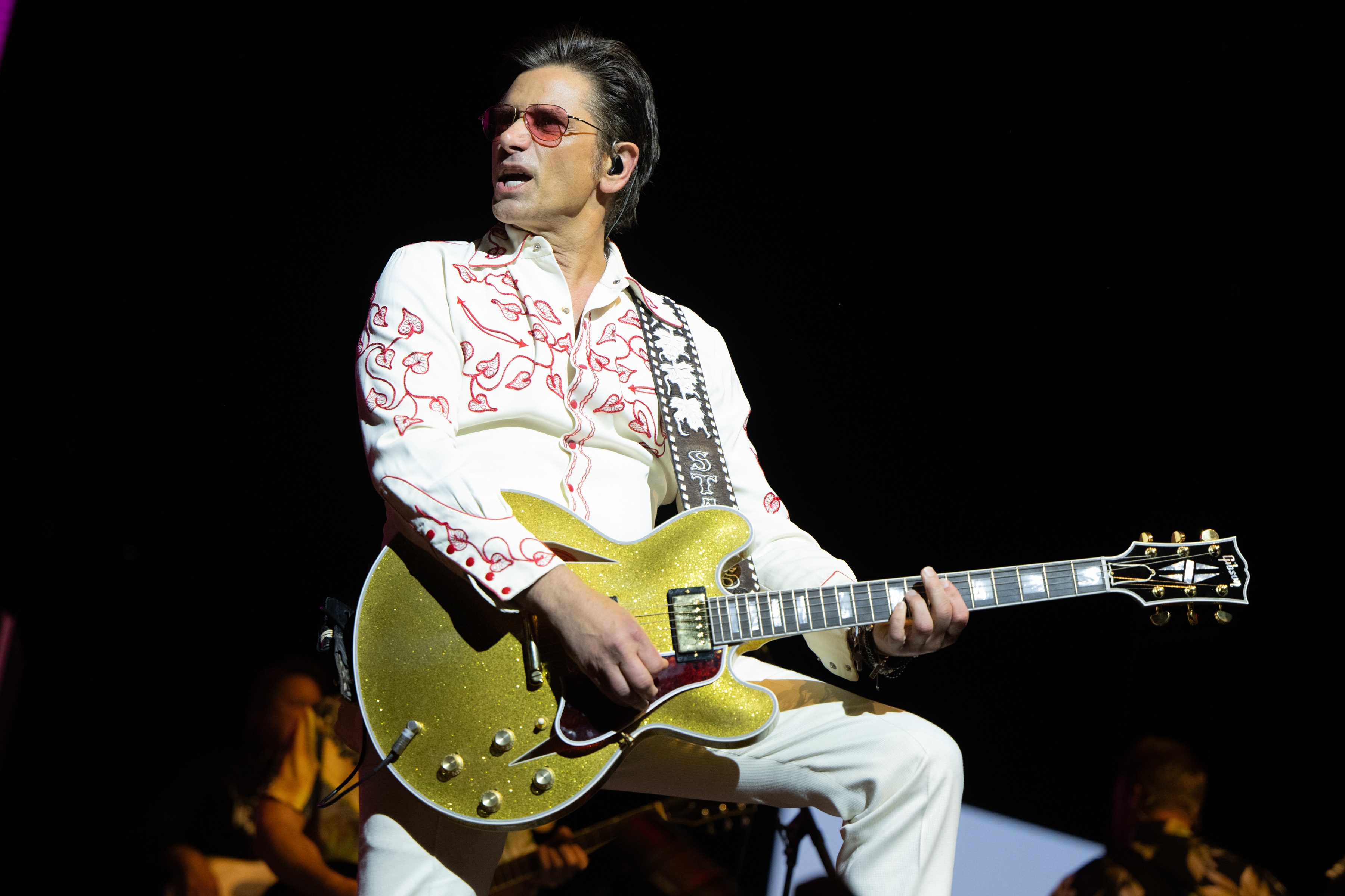 John Stamos performs on stage playing a gold guitar, wearing an embroidered white shirt and white pants