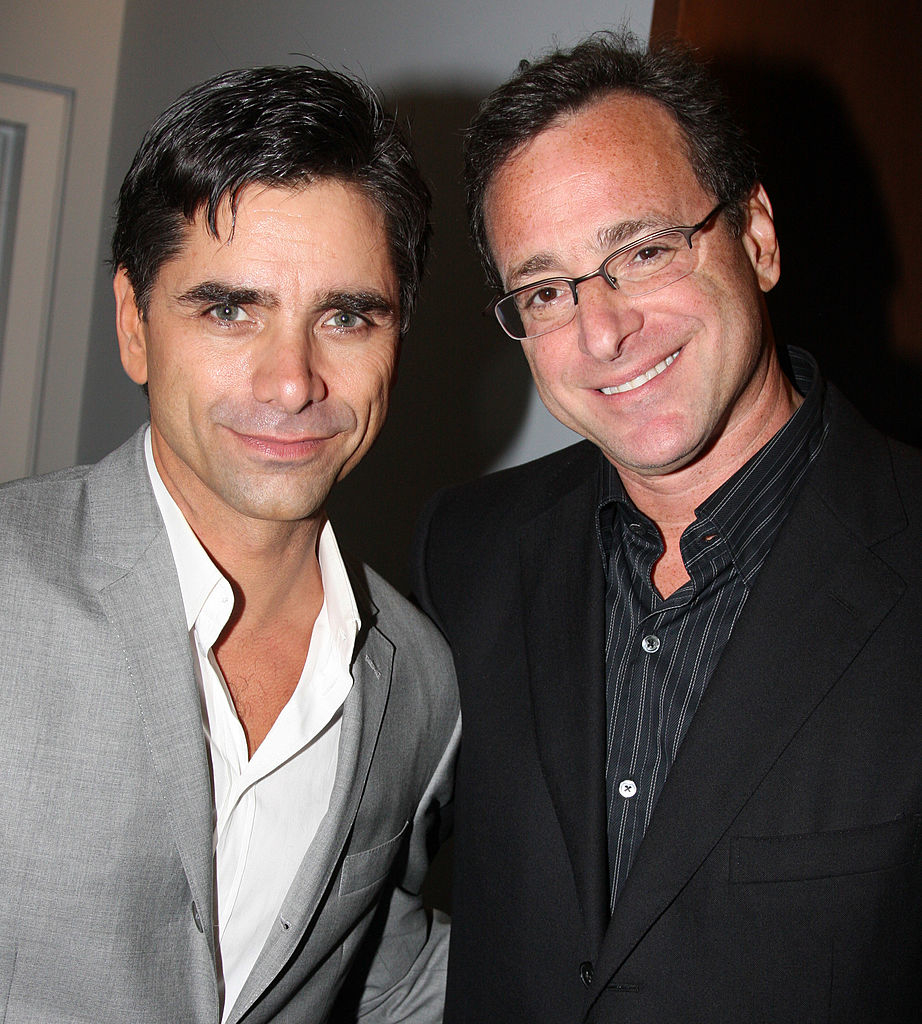 John Stamos in a gray suit and white shirt stands next to Bob Saget, who is wearing a black suit and dark striped shirt. Both are smiling at the camera