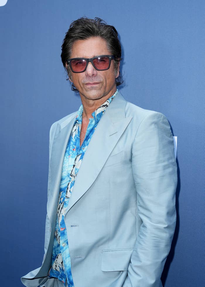 John Stamos poses on the red carpet in a light suit and patterned shirt, wearing sunglasses