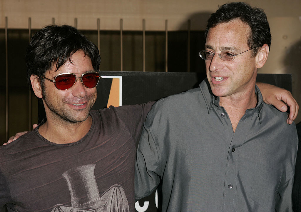 John Stamos and Bob Saget smile, standing side by side with arms around each other, at a casual outdoor event