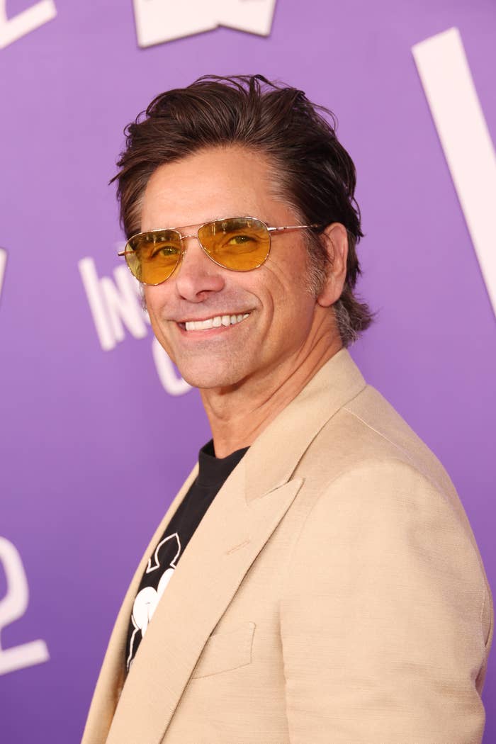 John Stamos, wearing a beige blazer and yellow sunglasses, smiles at a red carpet event