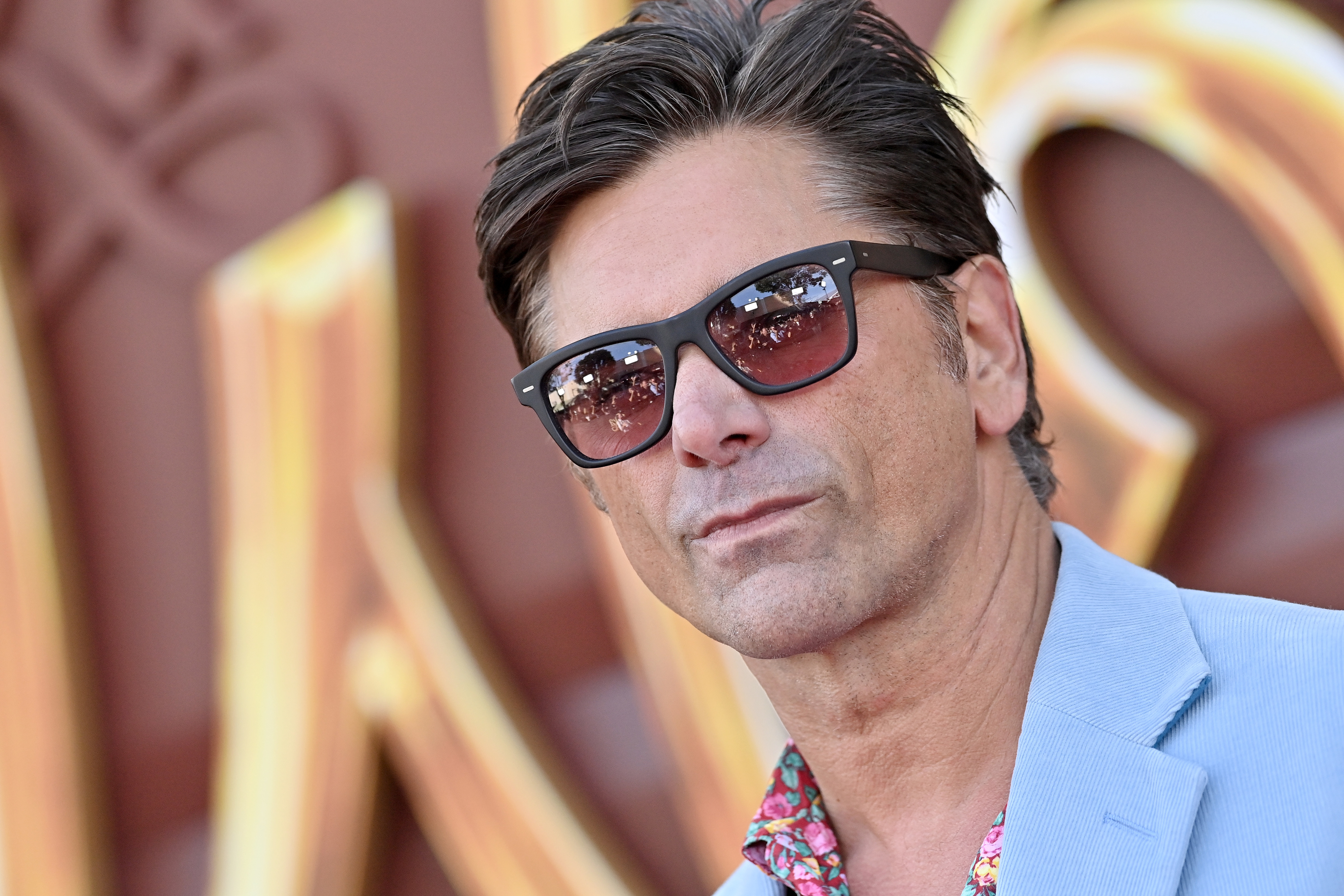 John Stamos wearing sunglasses, a light blazer, and a floral shirt, standing on a red carpet