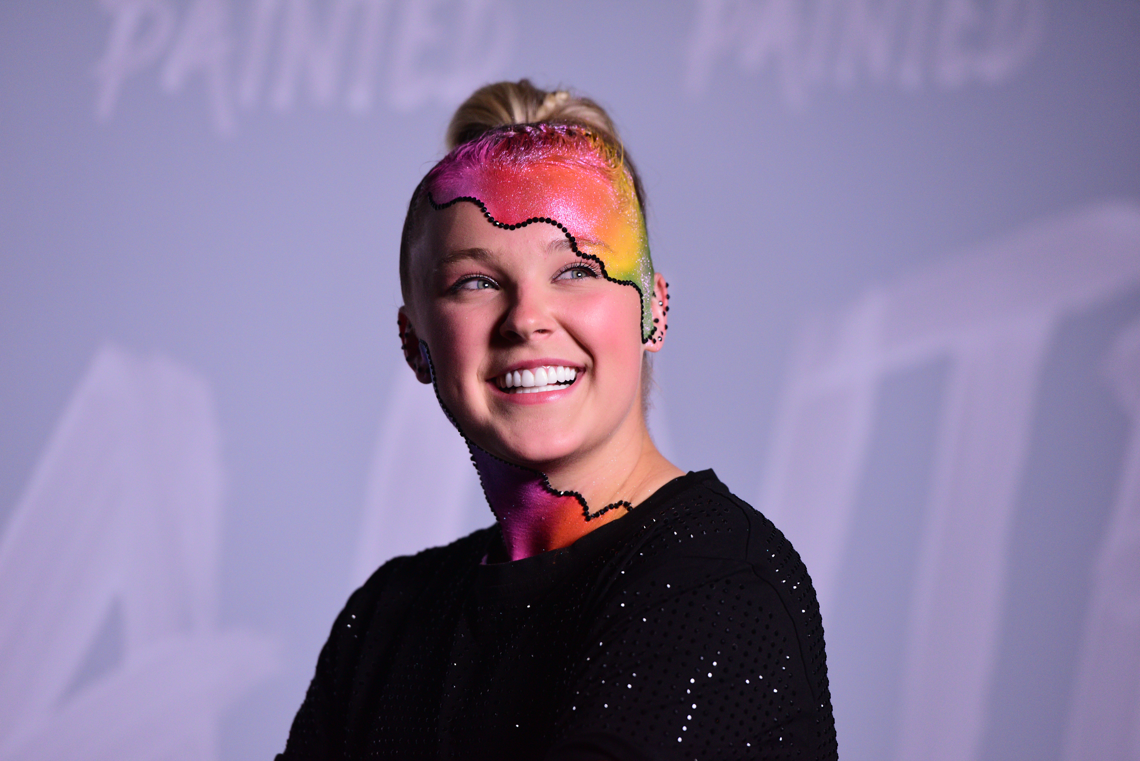 JoJo Siwa smiles while showcasing her vibrant, rainbow-colored face paint and wearing a black outfit at a celebrity event