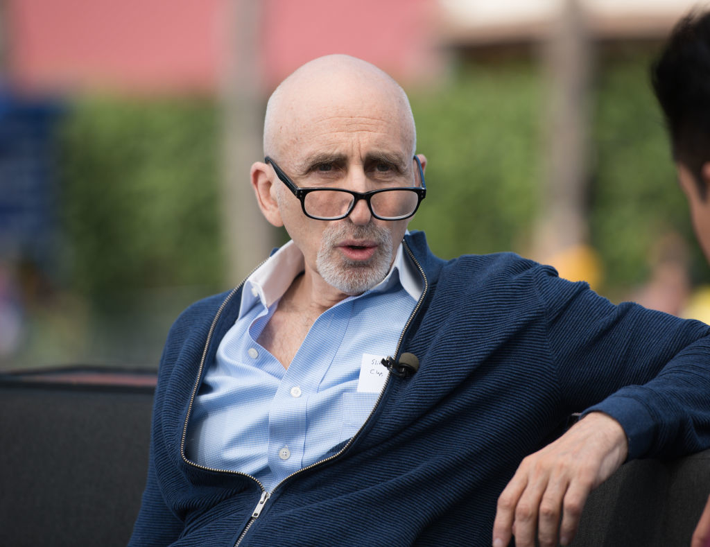 A bald man with a beard, wearing glasses and a blue zip-up sweater over a light blue shirt, is engaged in conversation outdoors