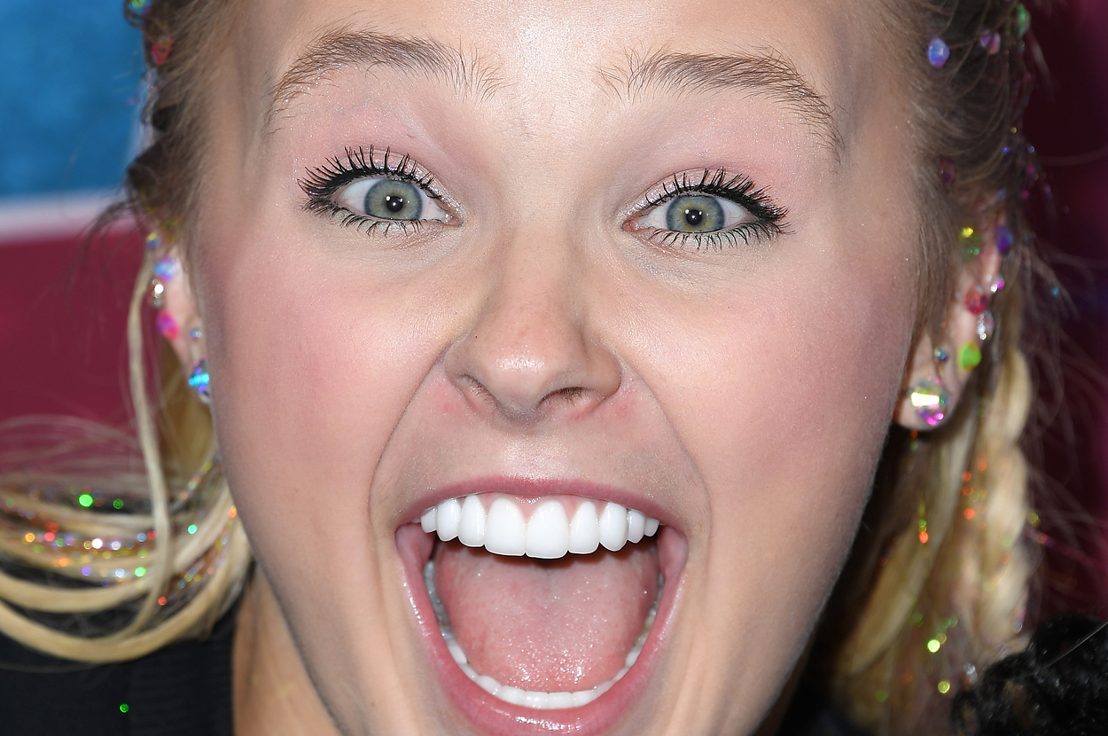 JoJo Siwa smiling on the red carpet with hair styled in braids and decorated with colorful gems