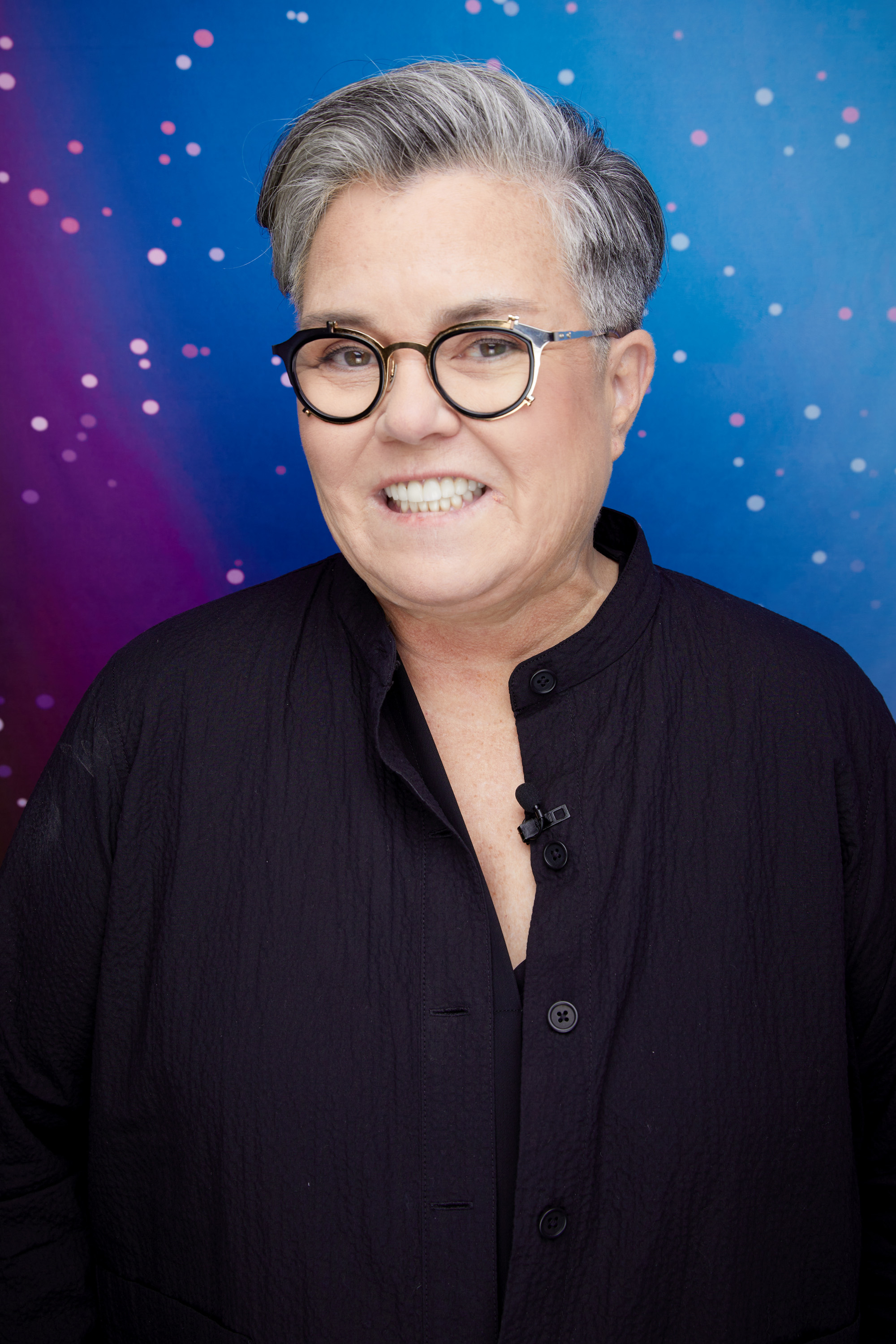 Rosie O'Donnell smiles while wearing a dark-buttoned top and black glasses, against a starry night background
