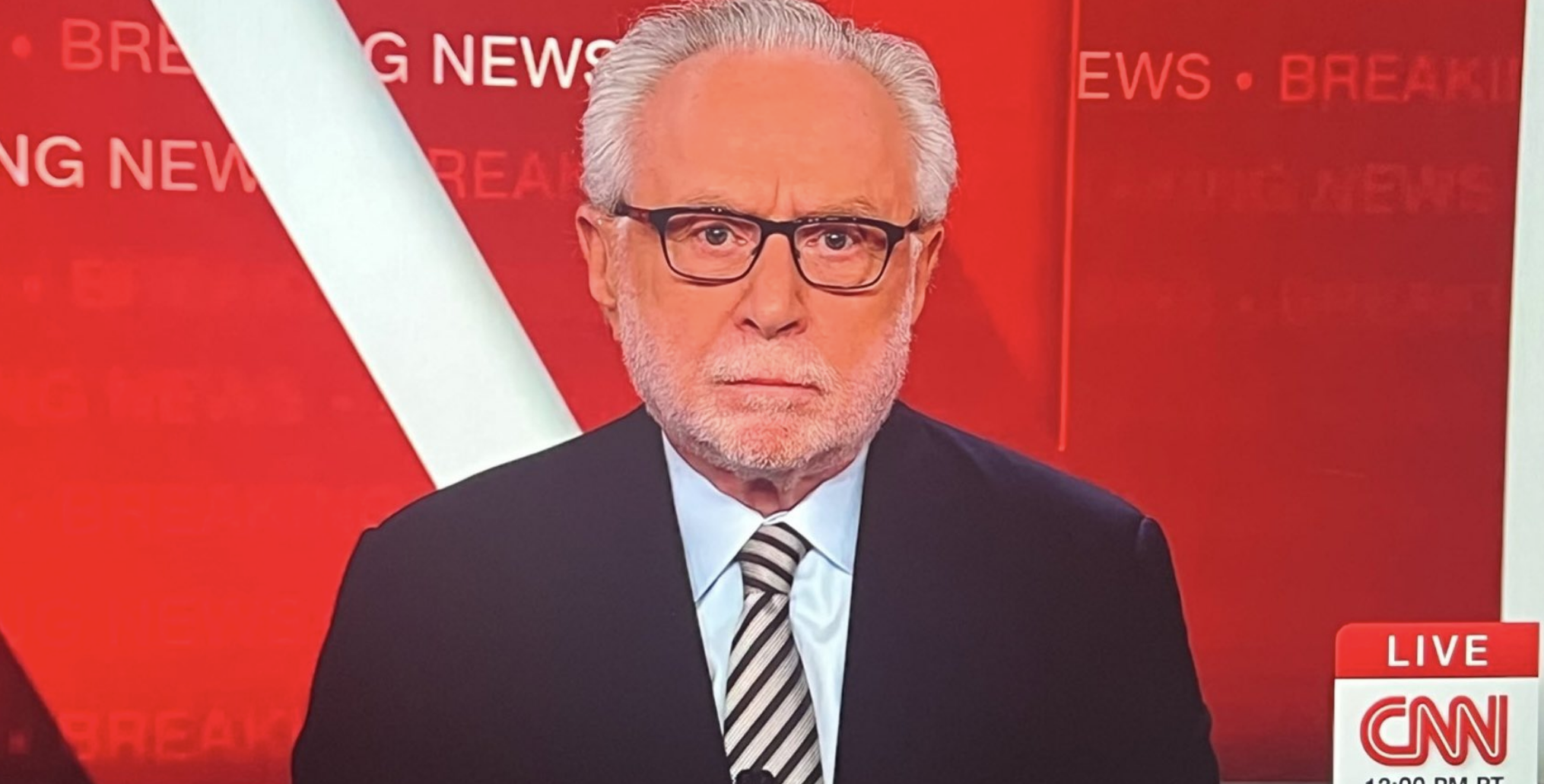 Wolf Blitzer on CNN breaking news set, wearing a suit and striped tie. The screen displays a live &quot;CNN Newsroom&quot; broadcast at 12:00 PM PT