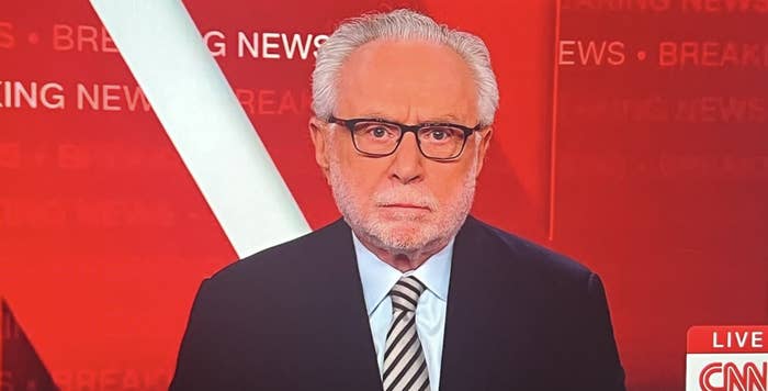 Wolf Blitzer on CNN breaking news set, wearing a suit and striped tie. The screen displays a live "CNN Newsroom" broadcast at 12:00 PM PT
