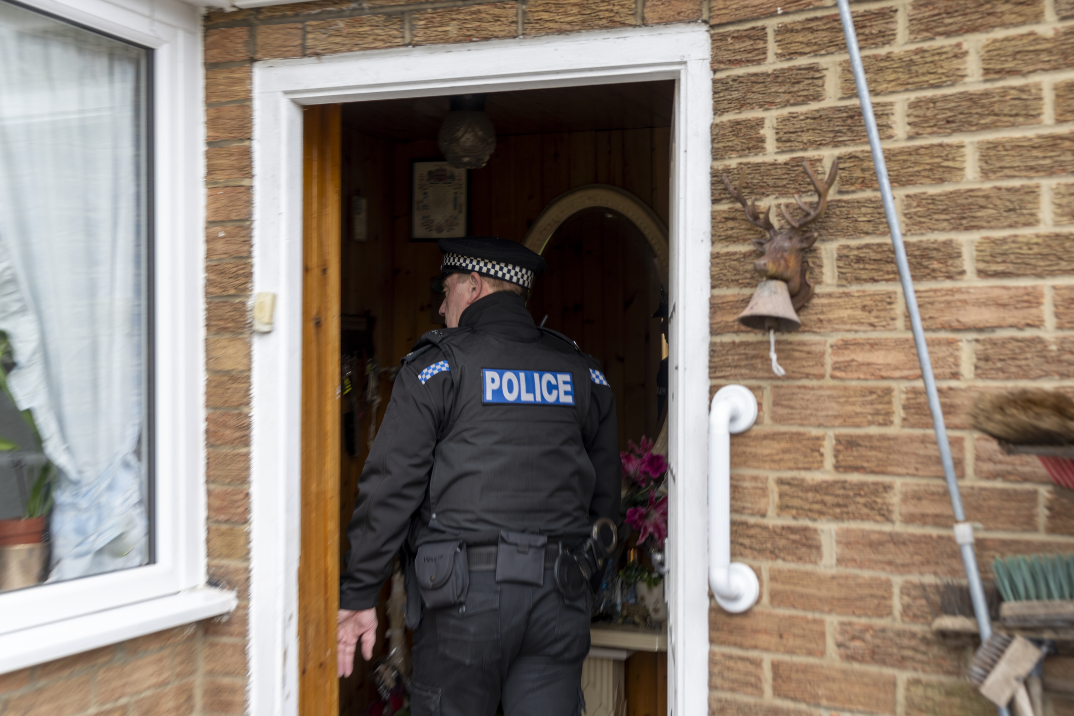 A police officer wearing a uniform enters a brick house through a front door. The setting appears to be a residential area
