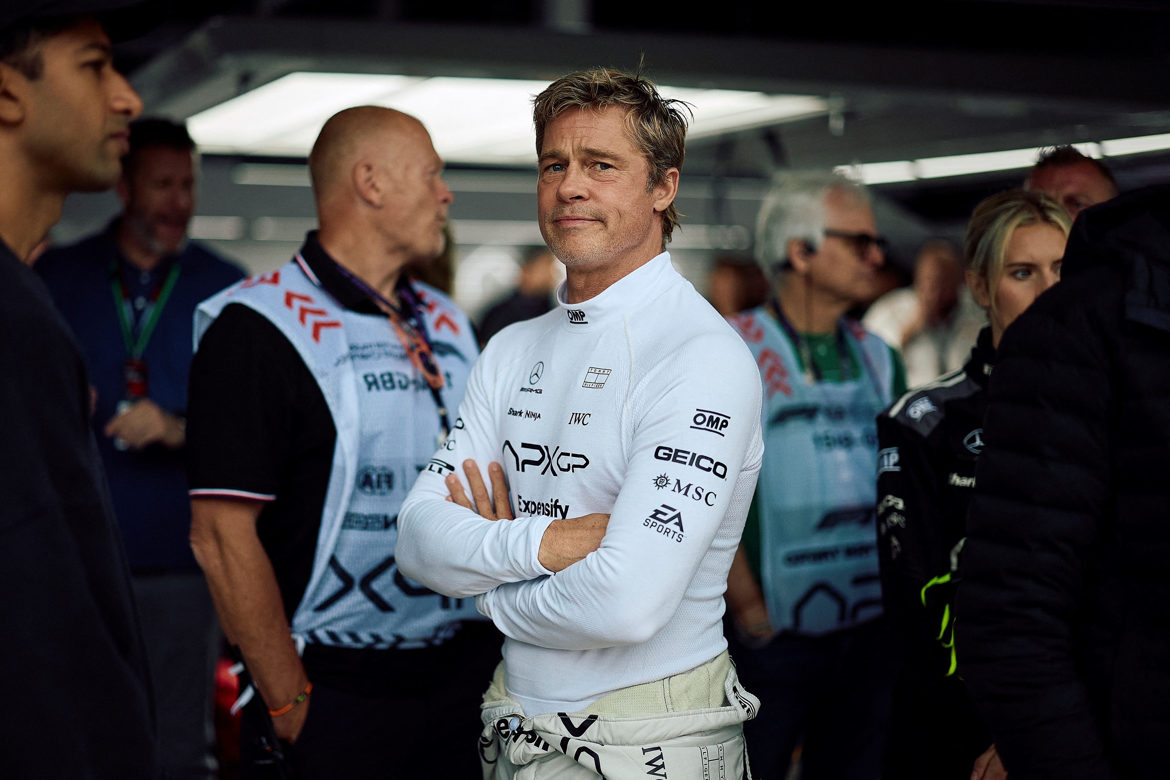 Brad Pitt in a racing suit with arms crossed, surrounded by team members and crew in a racing paddock