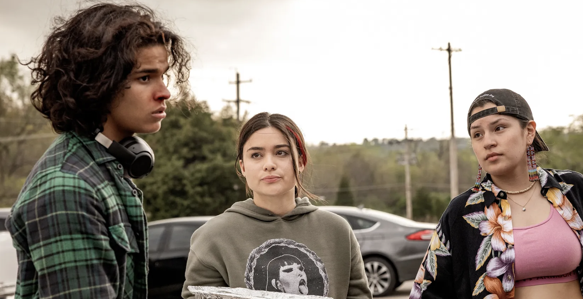 Devery Jacobs, Paulina Alexis, and Lane Factor stand outside, casually dressed; Devery in a hoodie, Paulina in a floral shirt and cap, Lane with headphones around his neck
