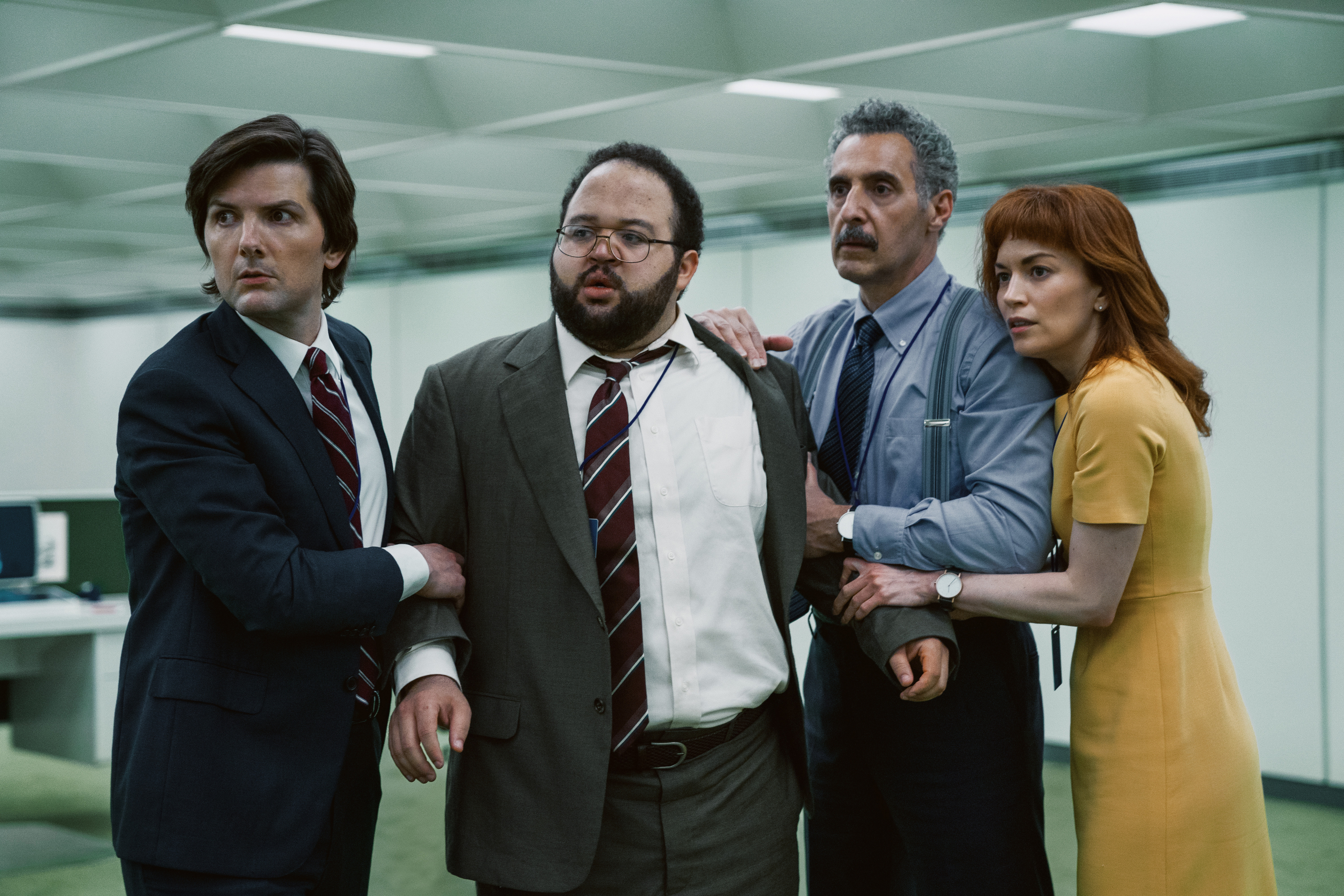 Adam Scott, Zach Cherry, John Turturro, and Britt Lower in business attire, standing in an office, looking concerned, and holding onto each other