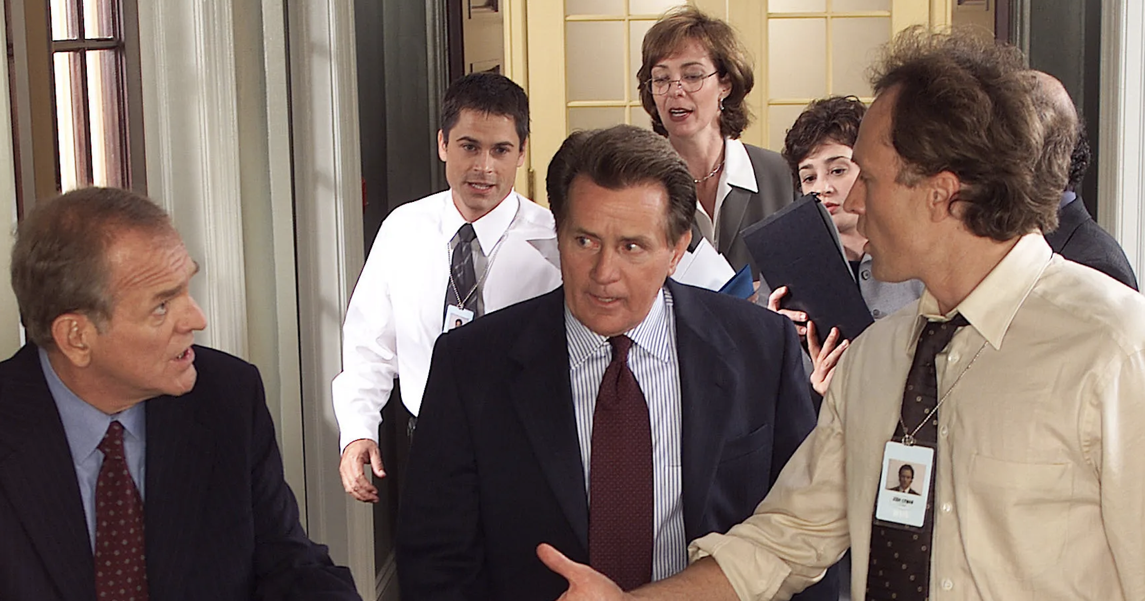 Martin Sheen, Rob Lowe, Bradley Whitford, Allison Janney, and Richard Schiff walking in a hallway, engaged in conversation, each dressed in professional attire