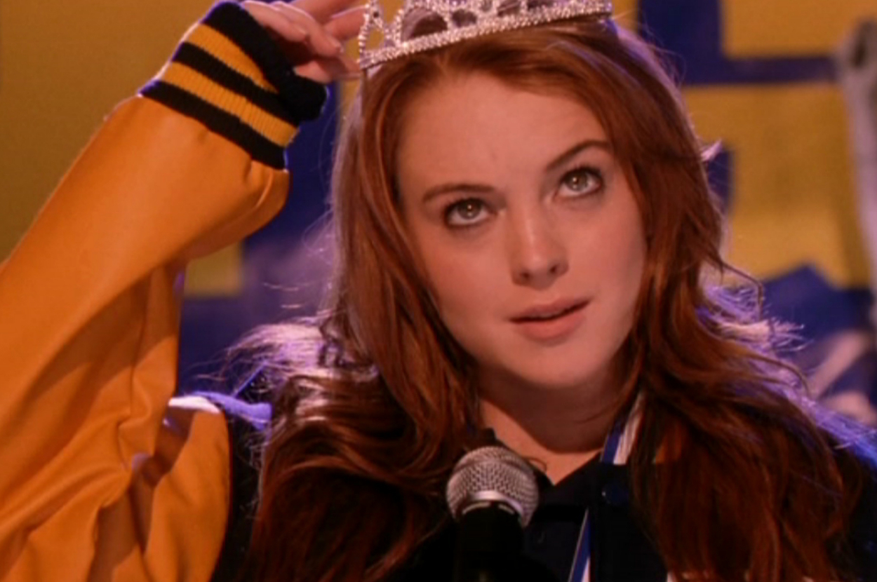Lindsay Lohan, in a scene from the movie "Mean Girls," is wearing a varsity jacket and holding a tiara above her head while speaking into a microphone
