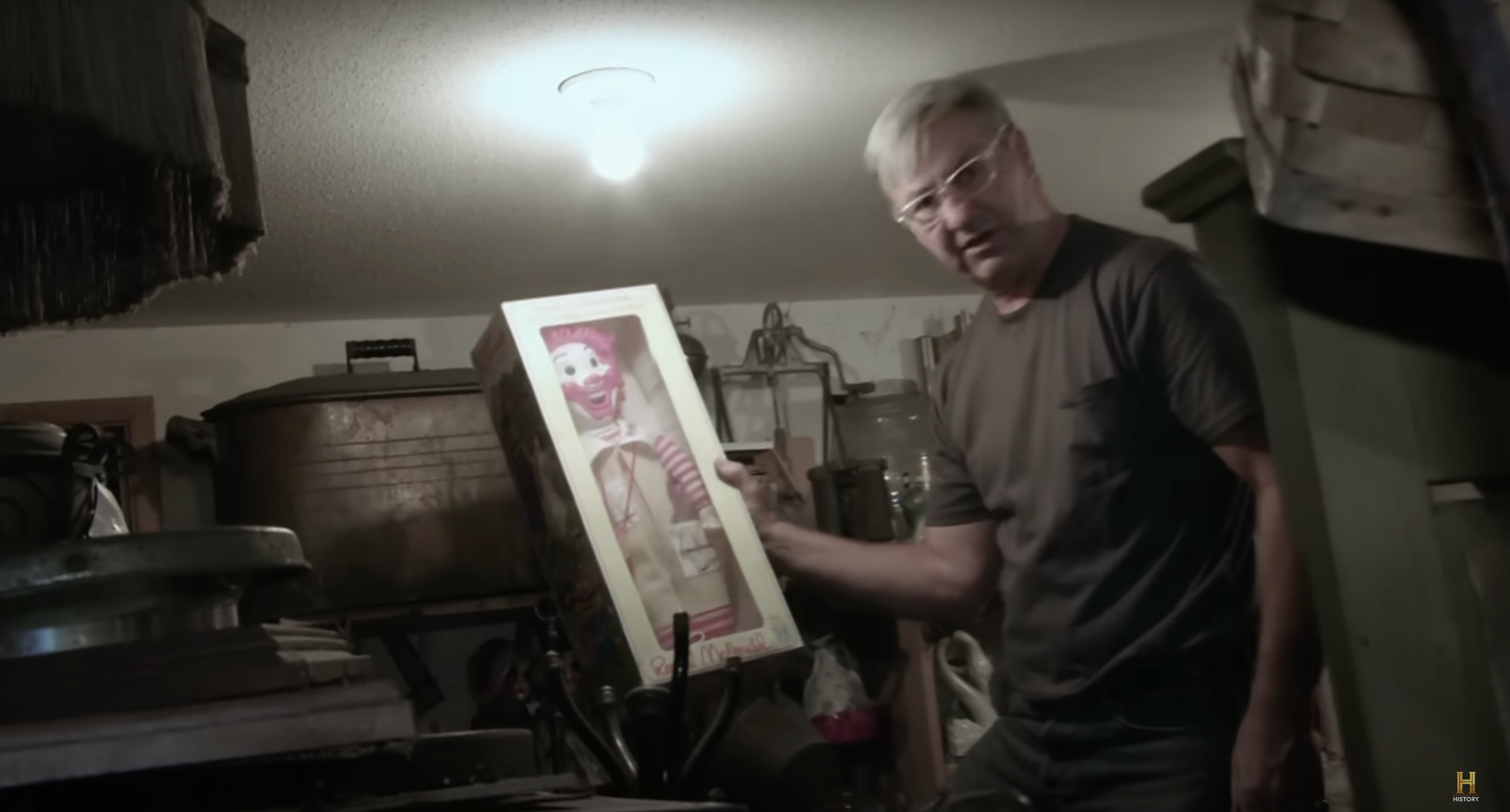 Man in a room holding a boxed clown doll, tilting the box towards the camera. The room has various antique items