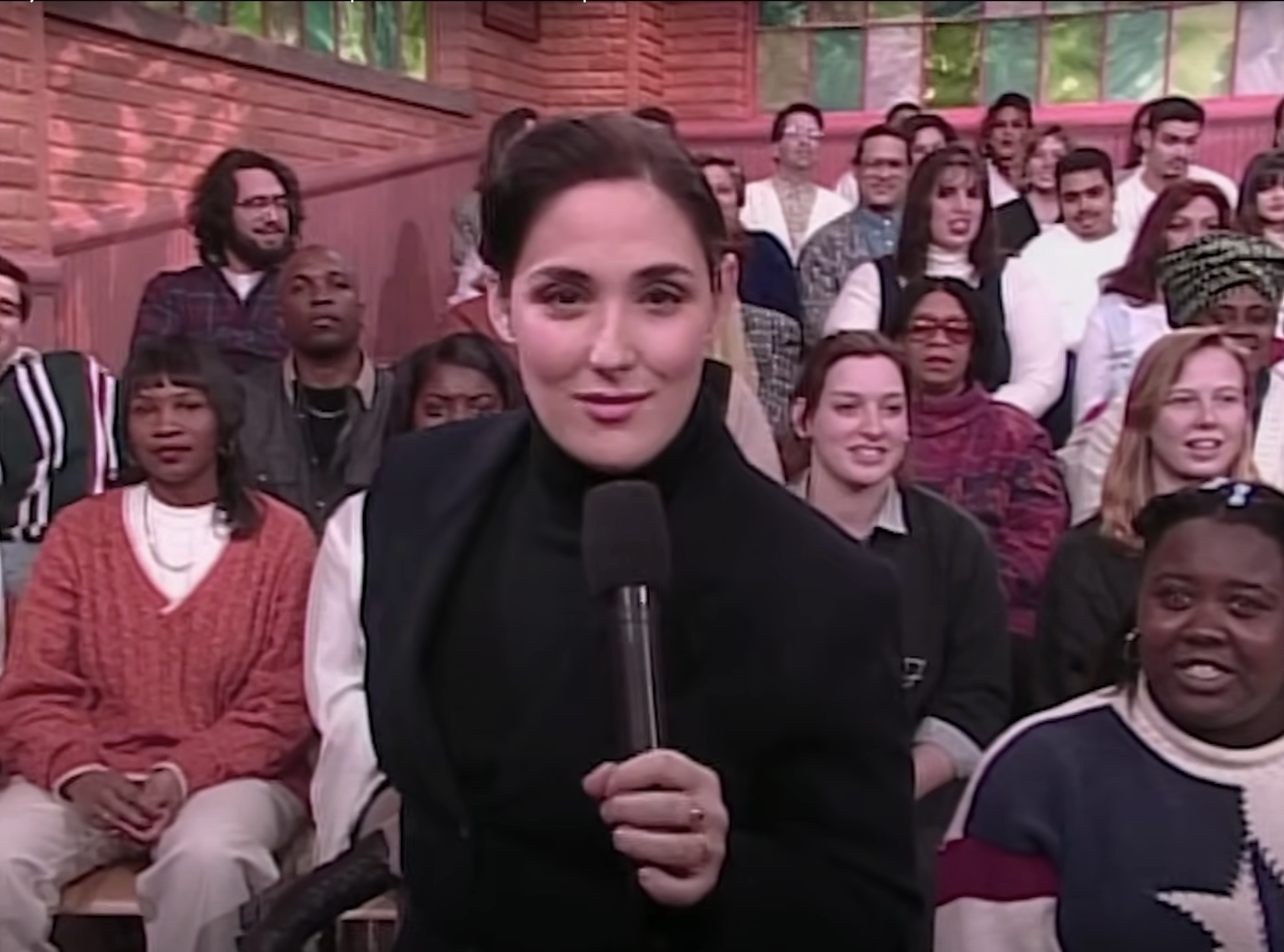 Ricki Lake stands with a microphone, engaging an audience sitting behind her, who appear excited and attentive