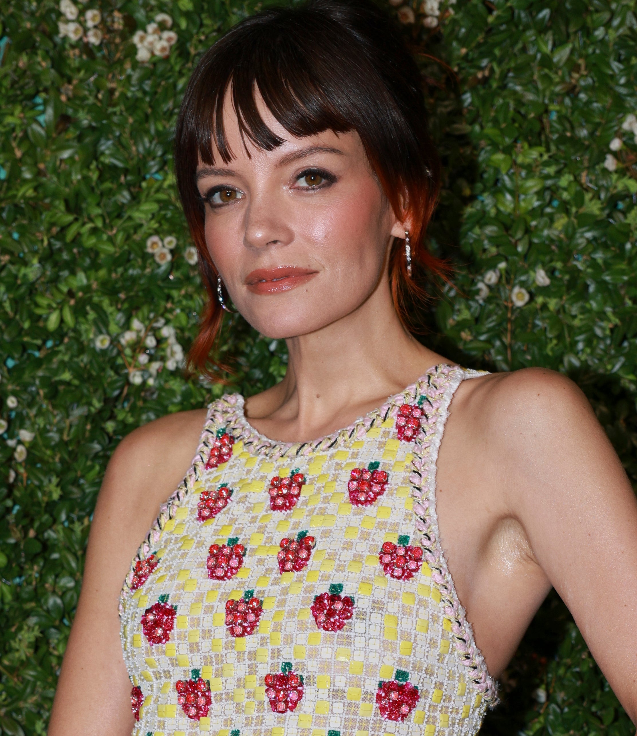 Lily Allen poses in front of a green foliage backdrop wearing a sleeveless dress with a floral pattern