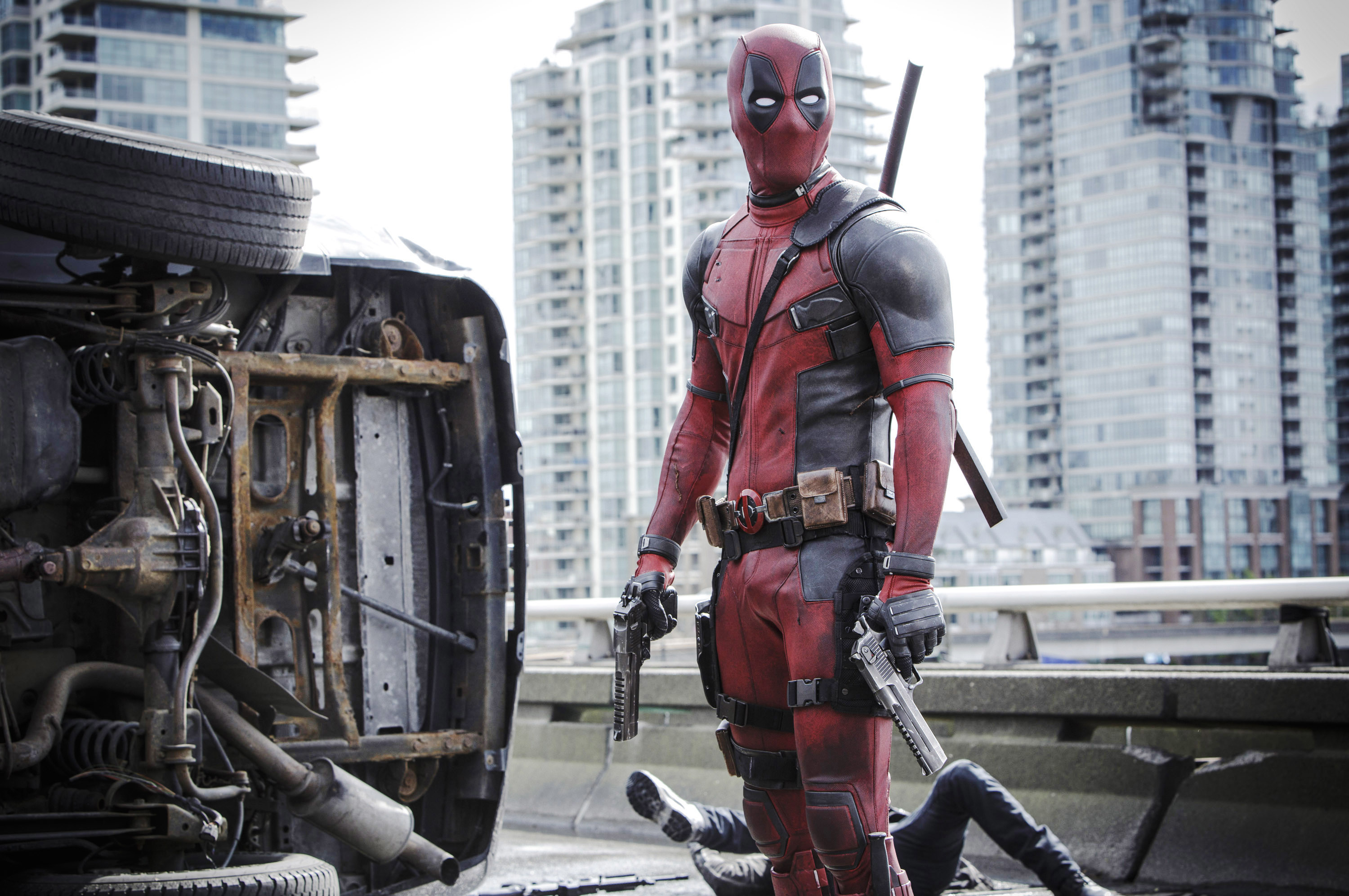 Deadpool in full costume stands on a city street holding two guns, with an overturned vehicle and buildings in the background