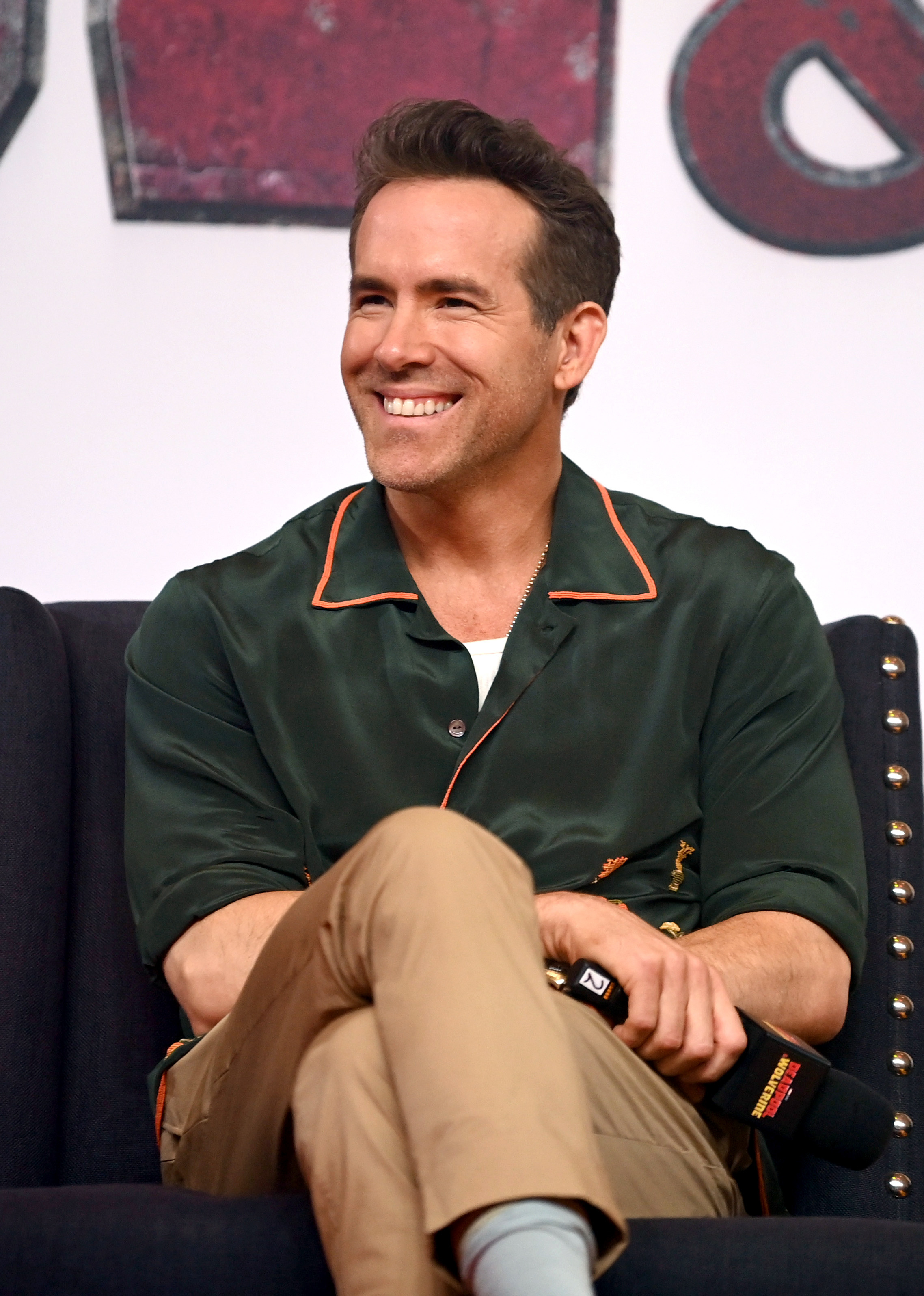 Ryan Reynolds smiling and seated during an interview, wearing a dark button-up shirt with orange trim and khaki pants, holding a microphone