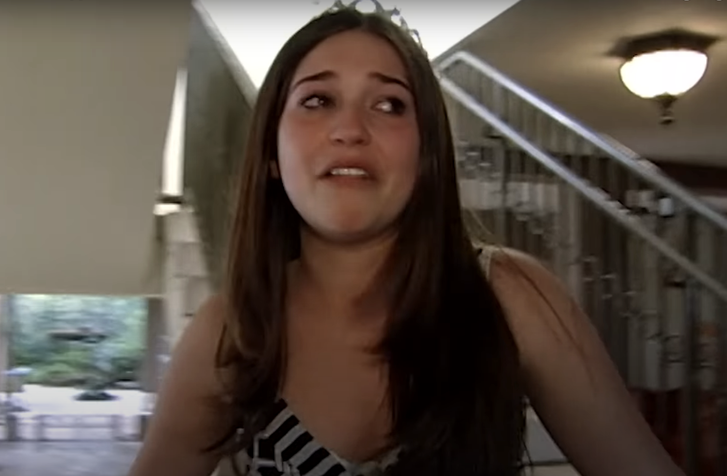 A teen girl with long hair wearing a sleeveless top and tiara stands by a staircase, appearing emotional