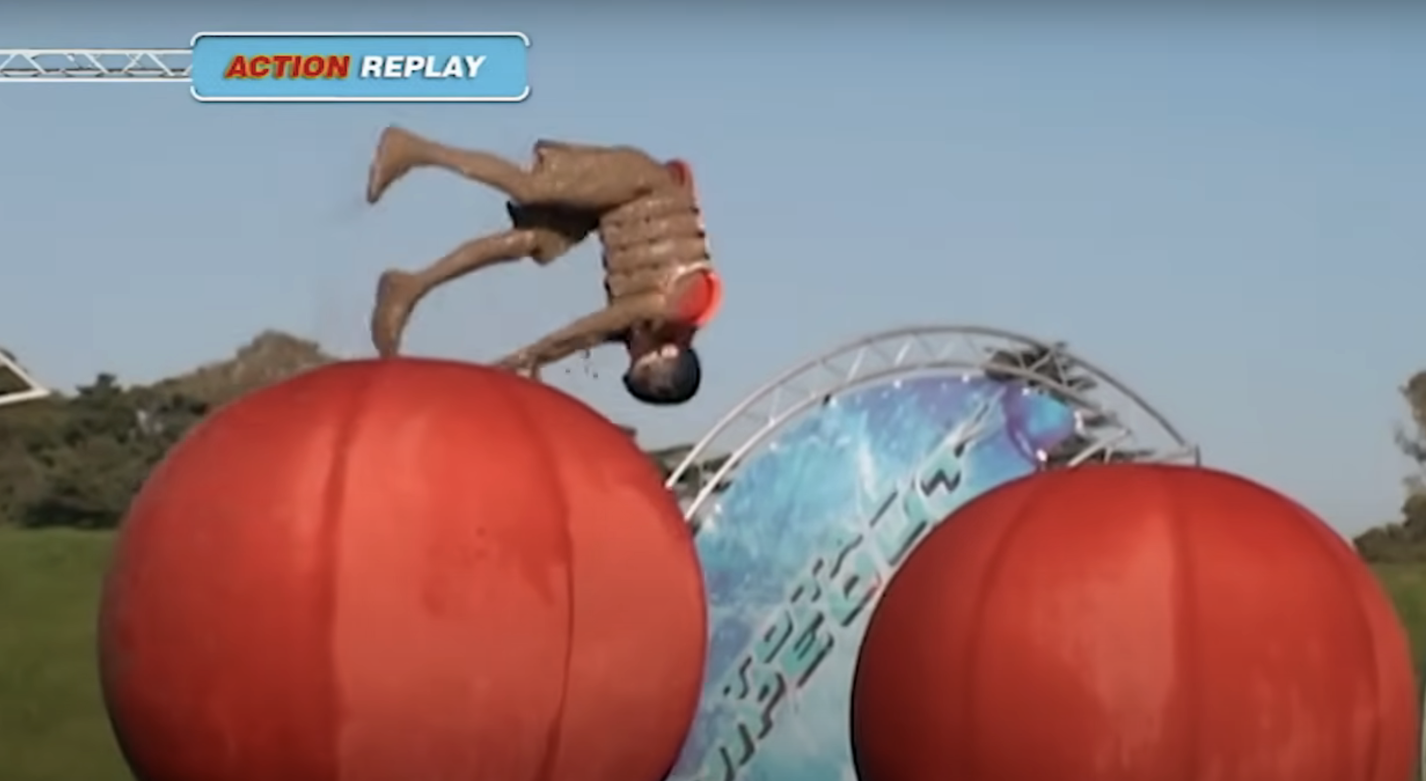 A mud-covered contestant from the TV show &quot;Wipeout&quot; flipping mid-air between two large balls during an obstacle course, shown in an action replay