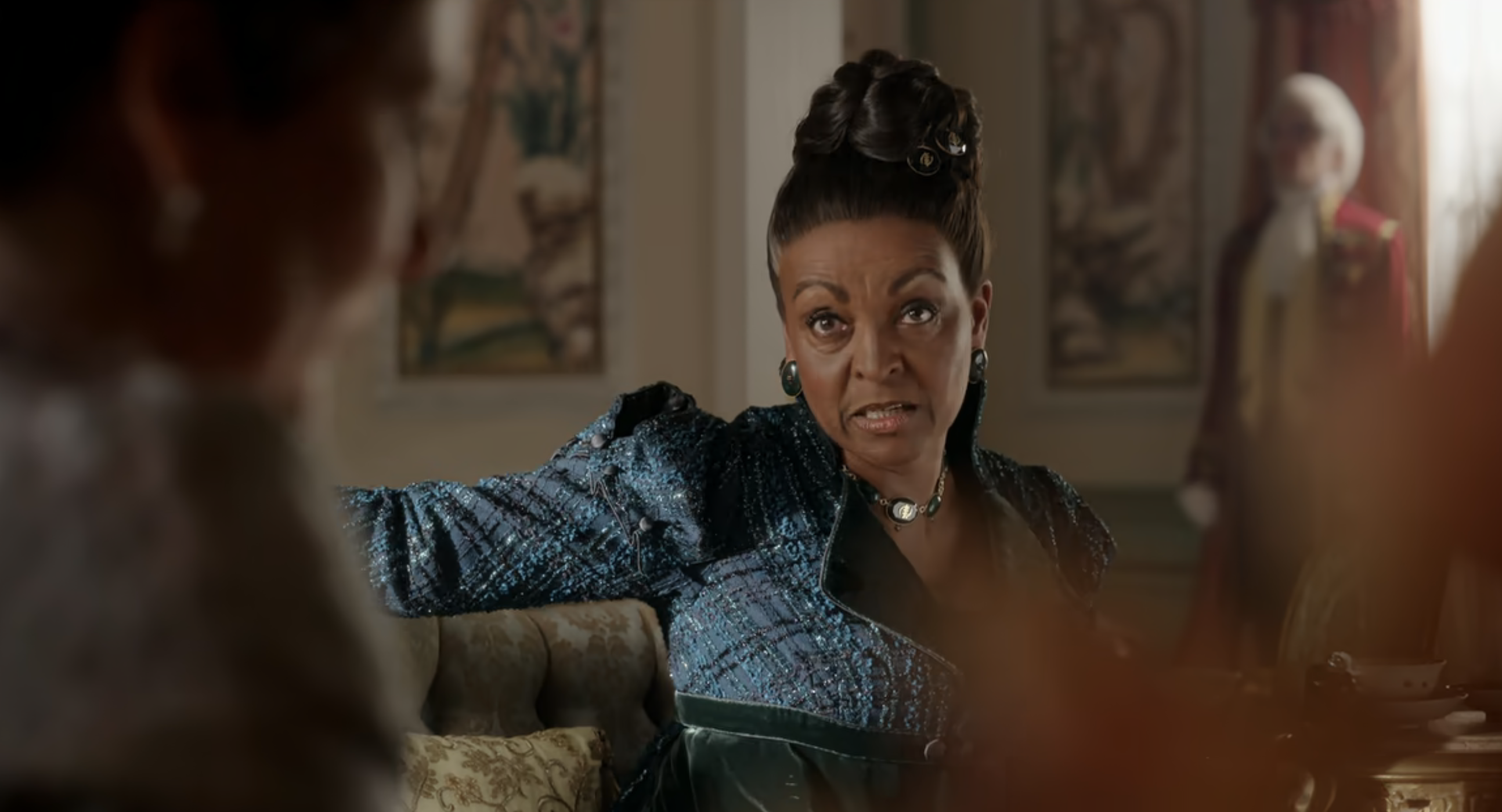 Adjoa Andoh in a period costume sits and gestures expressively in a room with historical decor, in a scene from a TV show or movie