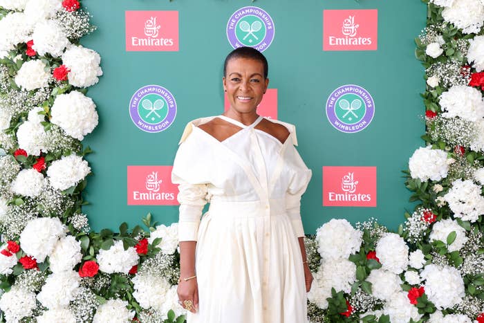 Adjoa Andoh stands on a floral-decorated backdrop at a Wimbledon event, wearing a stylish white dress with unique cutout details on the sleeves