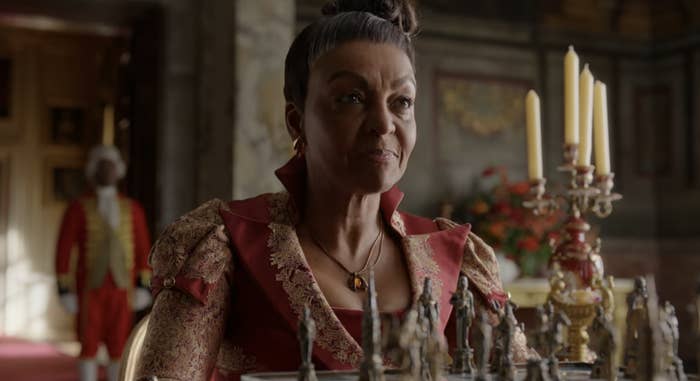 Adjoa Andoh dressed in an elaborate period costume sits at a table with chess pieces in a grand room, appearing thoughtful