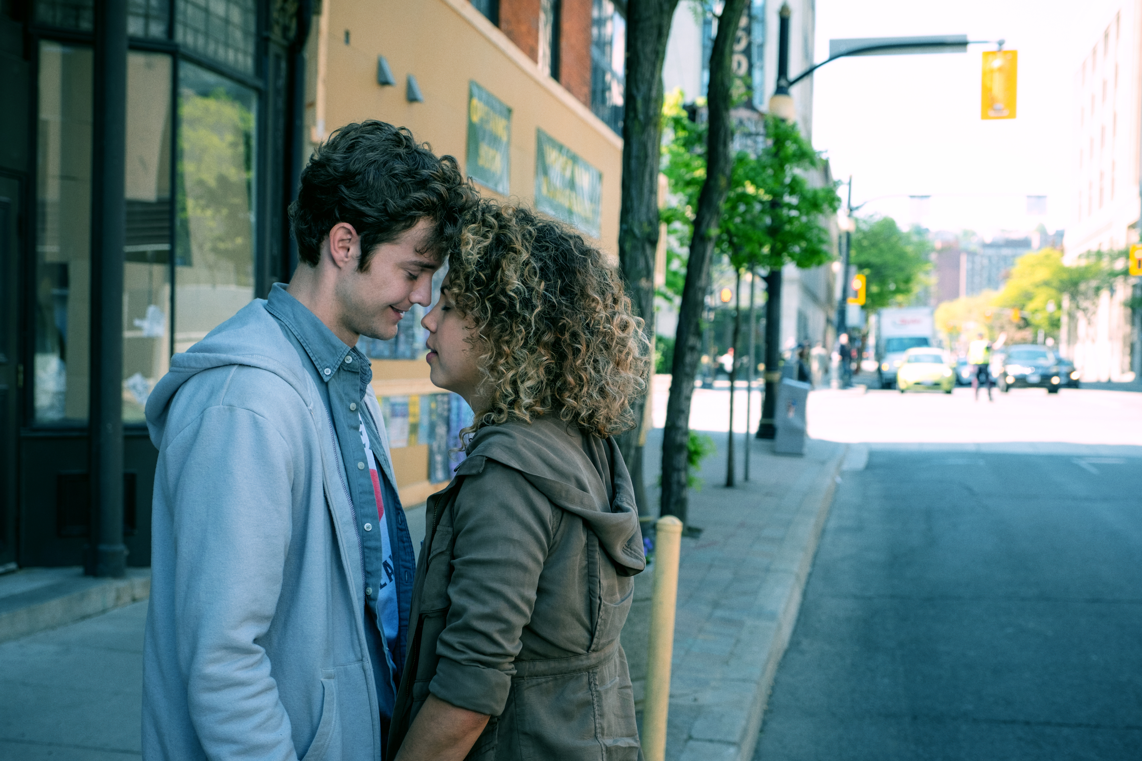 A man and a woman stand close, touching foreheads affectionately on a city sidewalk. They appear to be in a tender, intimate moment