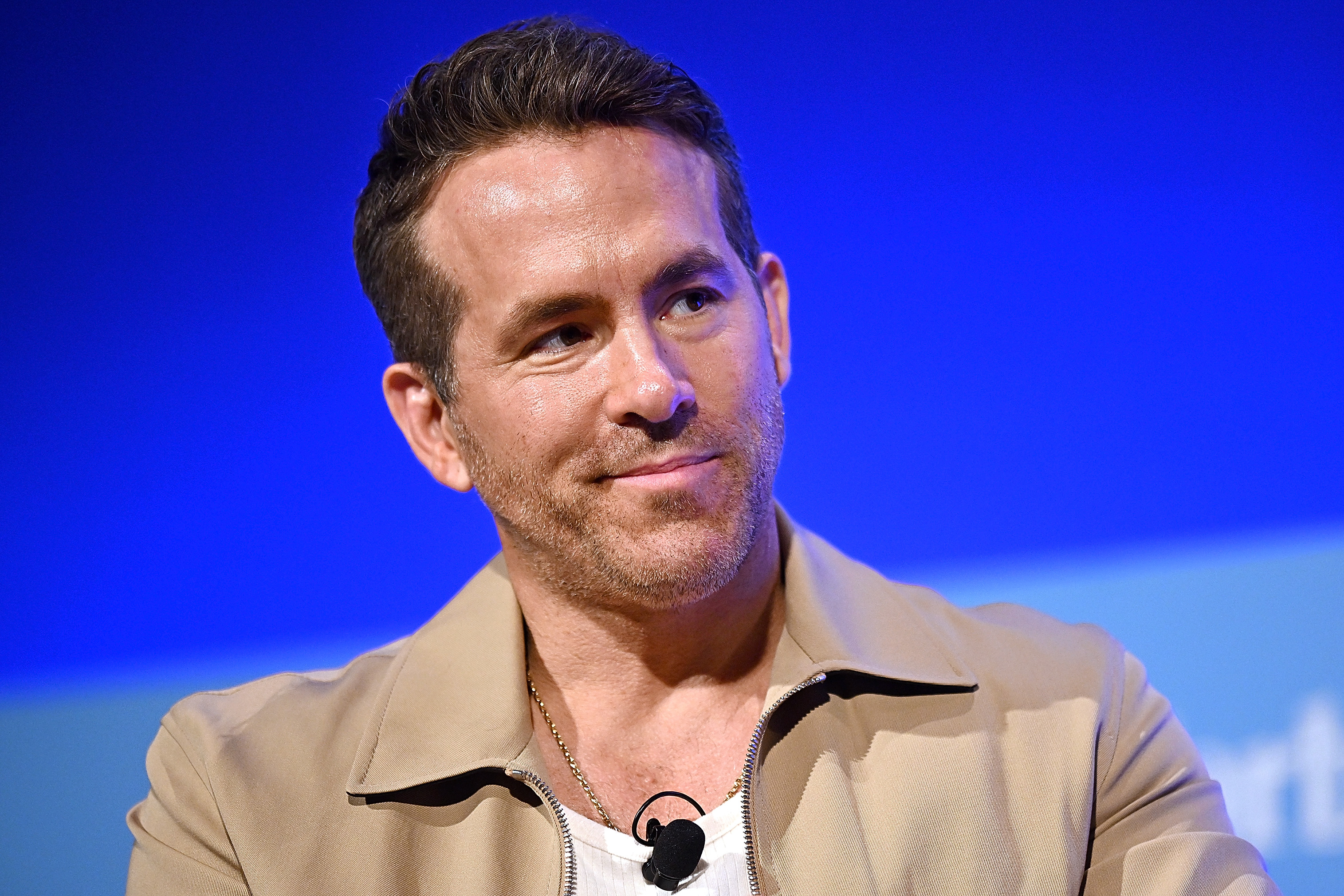 Ryan Reynolds smiles while wearing a beige jacket over a casual shirt during a speaking event