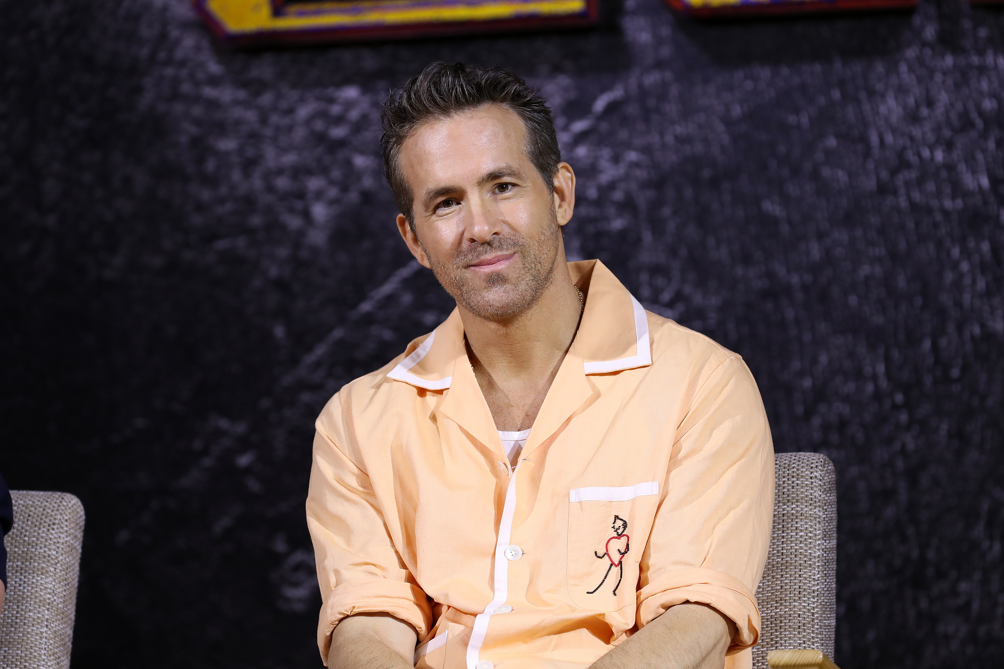 Ryan Reynolds is sitting and wearing a casual button-up shirt with a small embroidered design on the chest