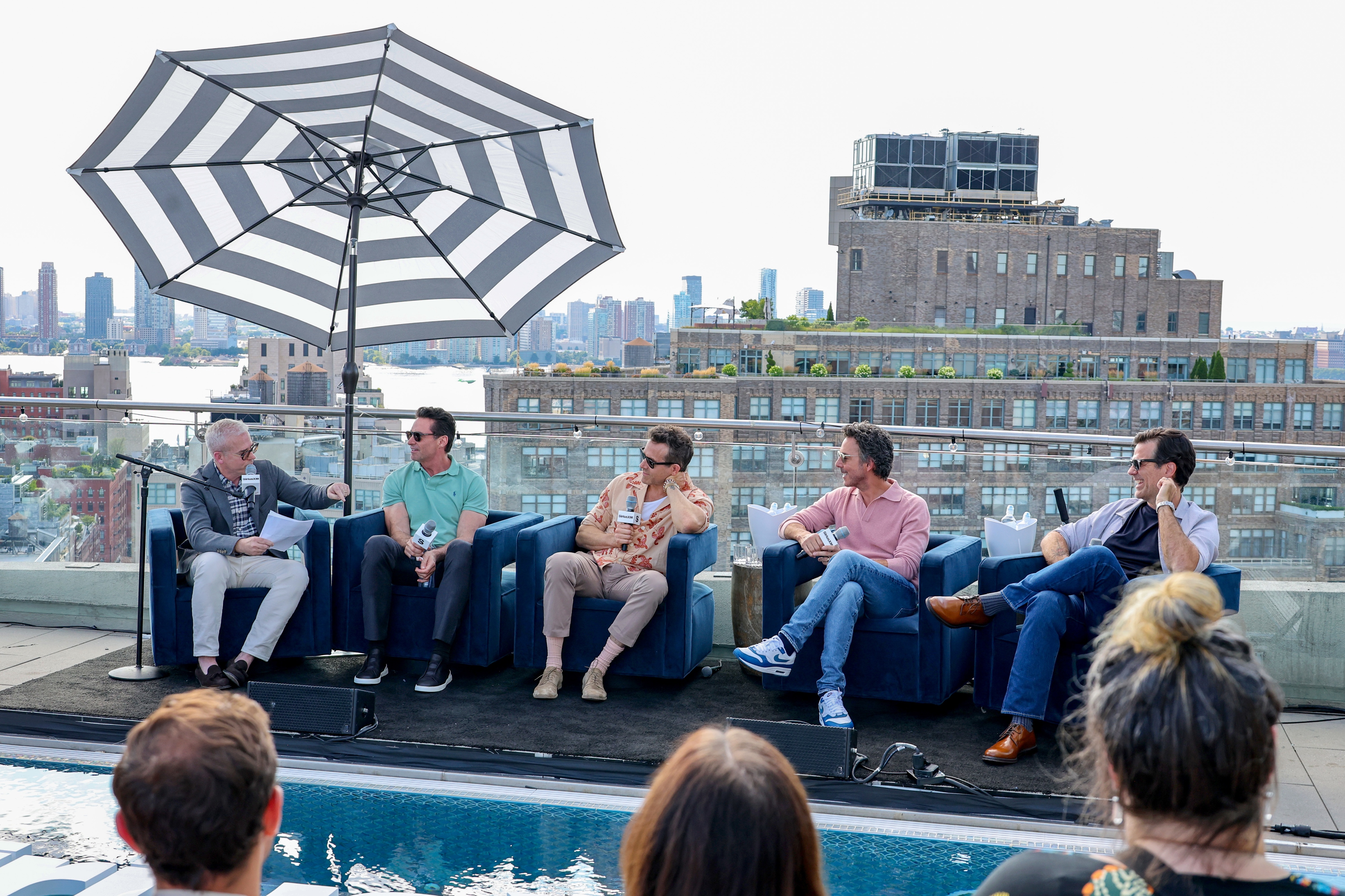 Panel discussion on a rooftop with a city skyline in the background. Four men seated under a striped umbrella, with one person moderating