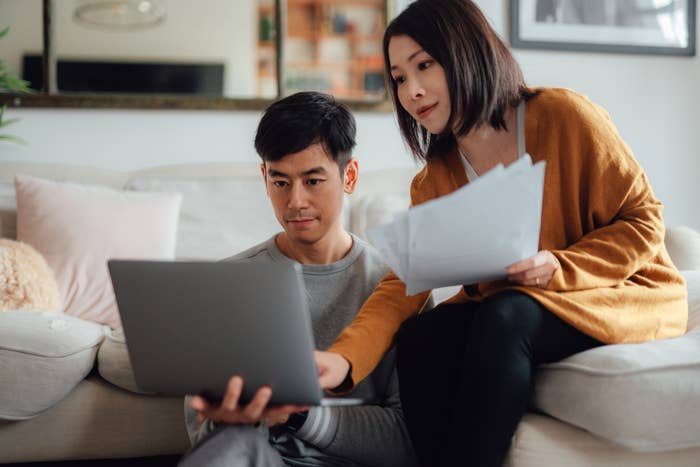 A couple is sitting on a couch, looking intently at a laptop while the woman holds documents