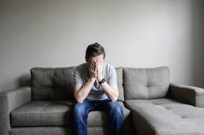 A man sits on a couch with his head in his hands, expressing distress