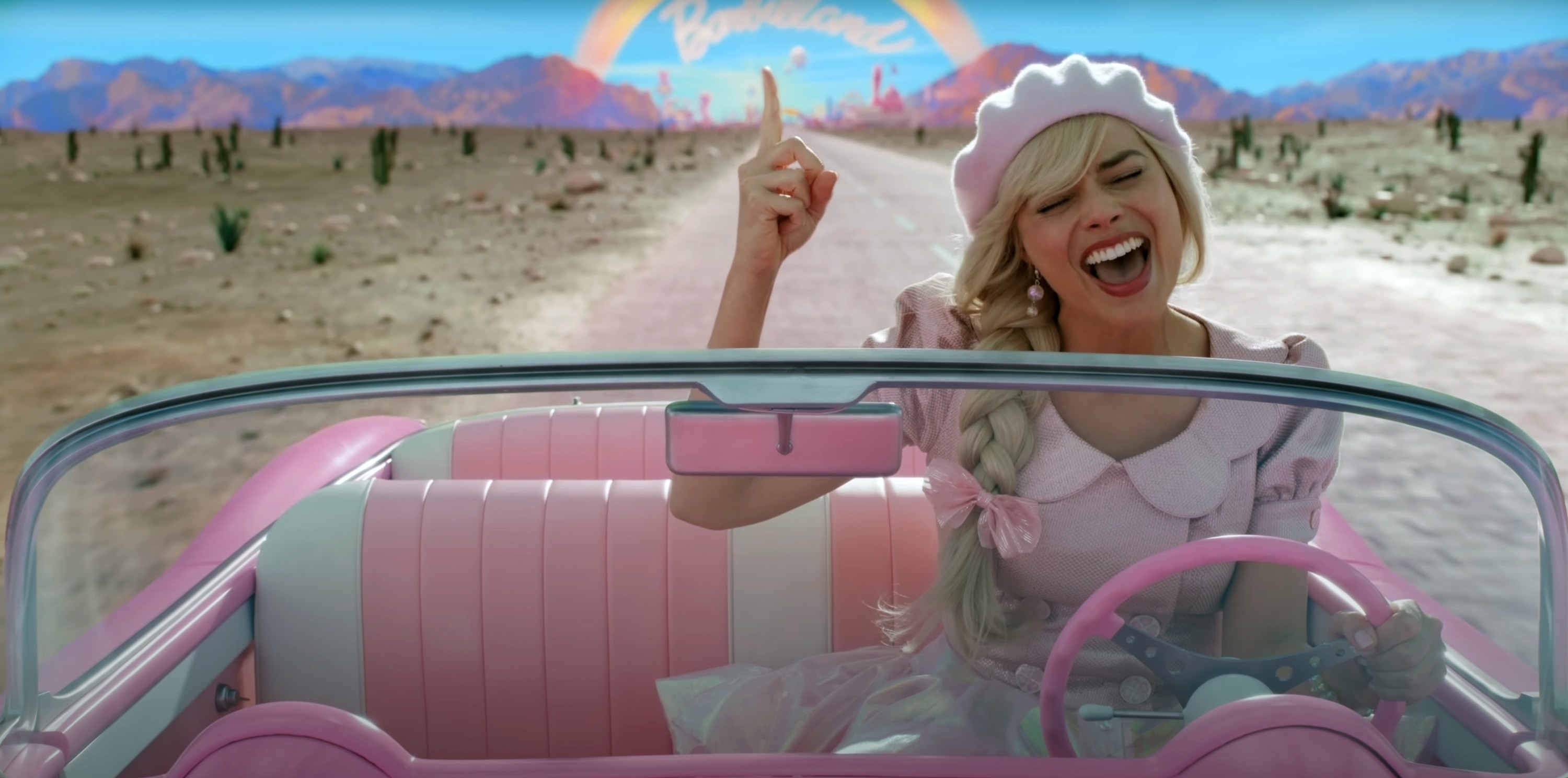 Barbie in a pink convertible, wearing a pink beret and dress, smiles and points up while driving through a desert towards a bright horizon that reads &quot;Barbieland.&quot;
