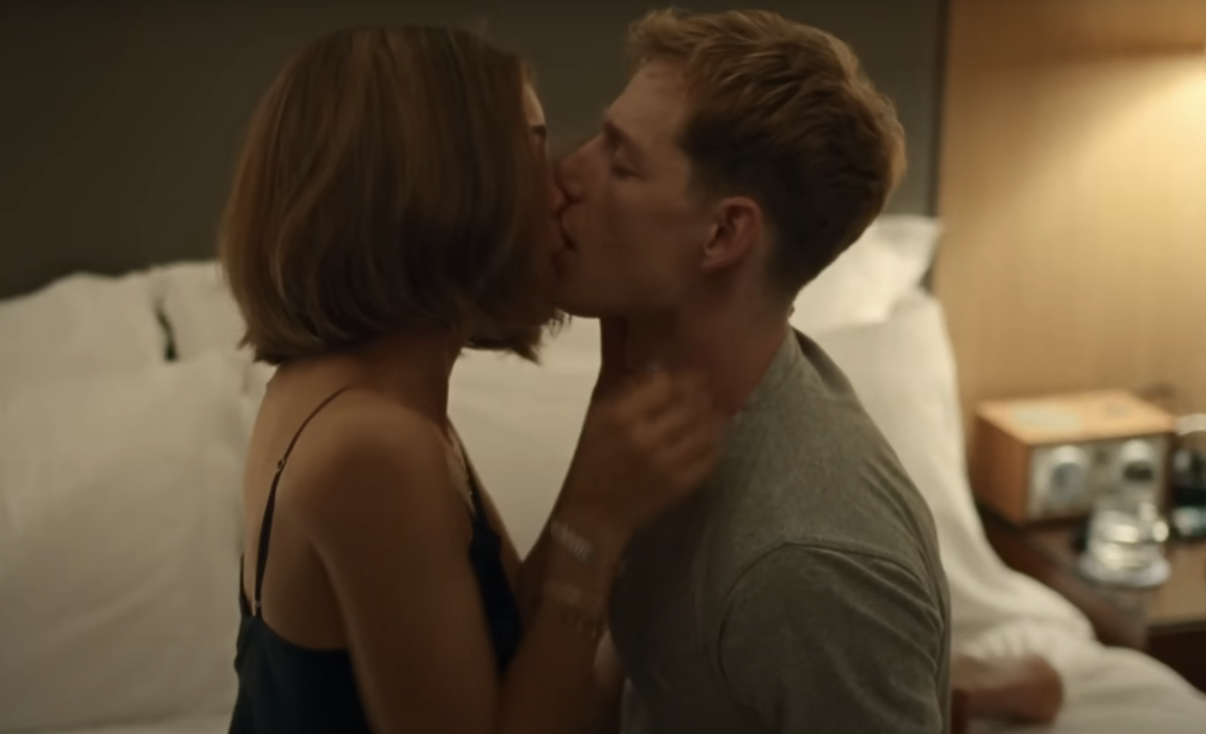 A woman and man share a passionate kiss while seated on a bed in a dimly-lit room