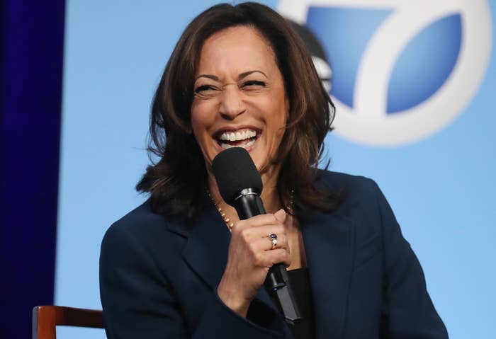 Kamala Harris holding a microphone, laughing while speaking at an event. She is wearing a dark blazer