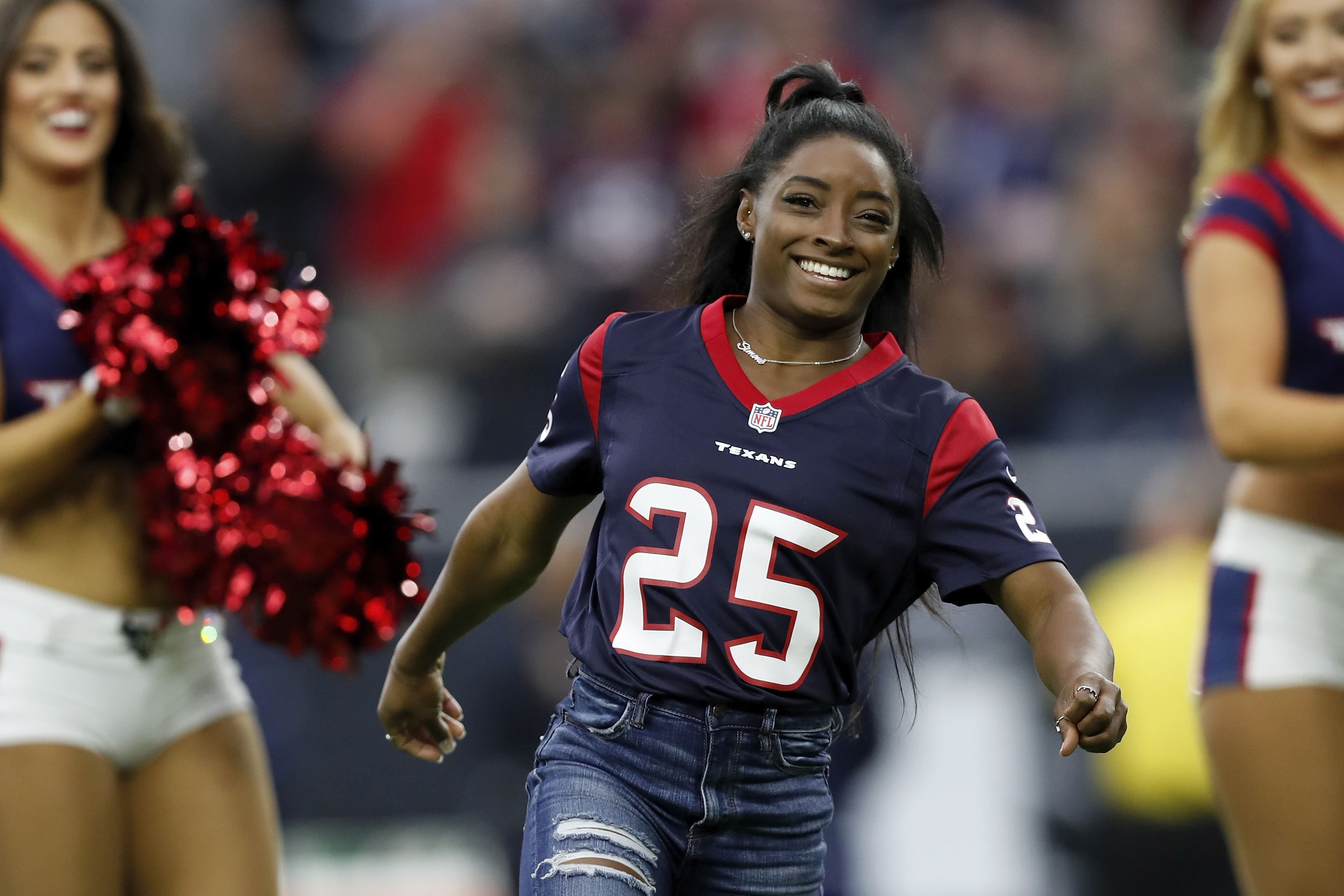 Simone Biles, wearing a Houston Texans jersey, smiles and runs on a football field with cheerleaders in the background