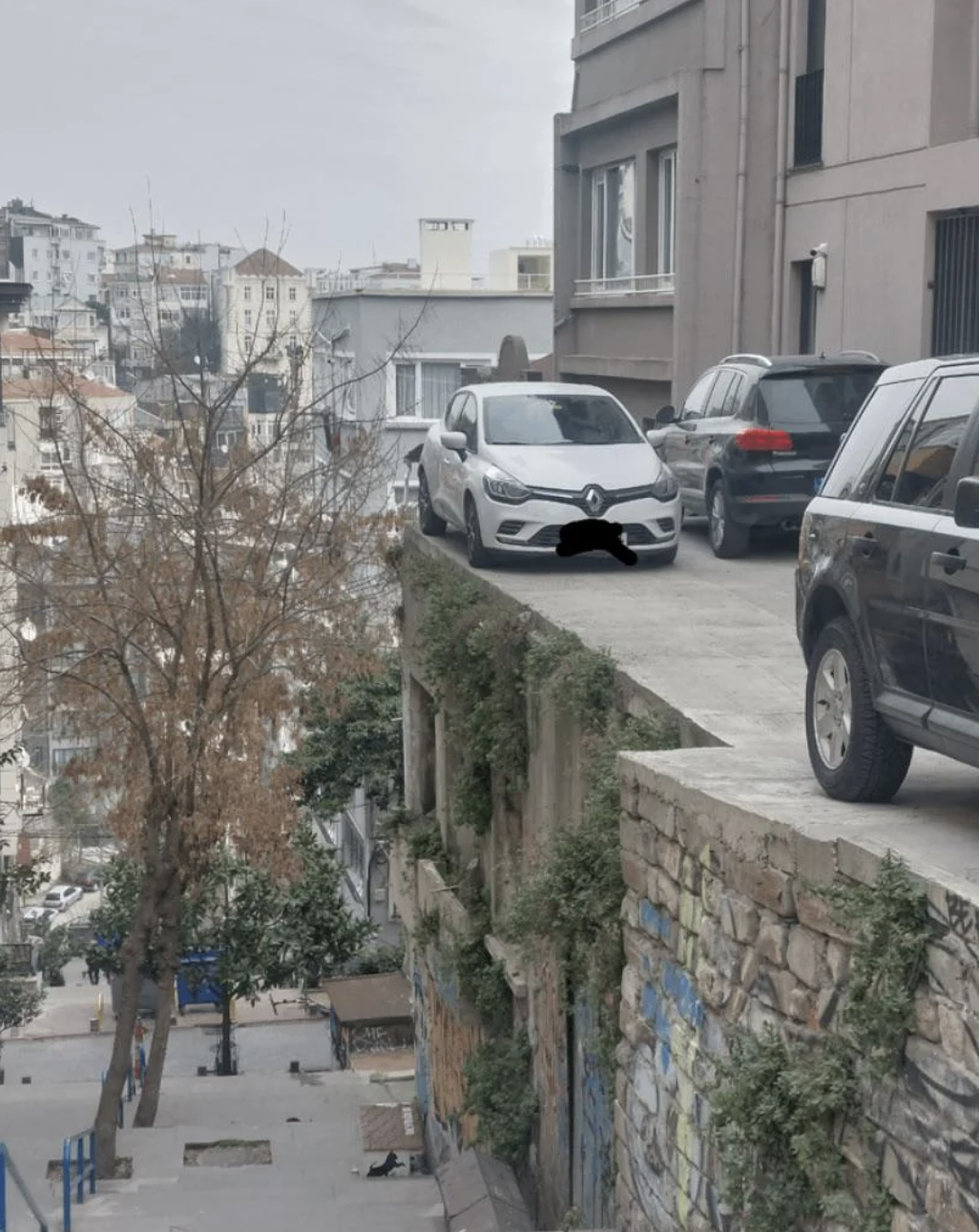 Cars parked on the edge of a rooftop-like structure in an urban area, creating a perilous scene. Pedestrians and graffiti can be seen below