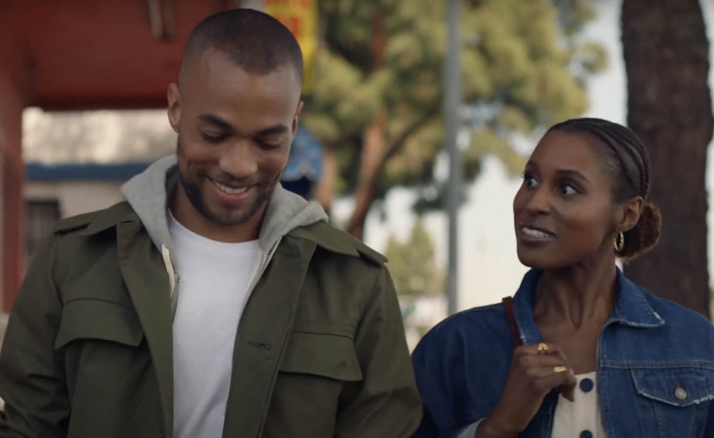 Two people walk outdoors; the man wears a jacket over a hoodie and shirt, while the woman wears a jean jacket