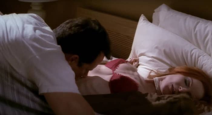 A man is kissing a red-haired woman’s neck as she lies in bed with her eyes closed, wearing a pearl necklace. Both people are fully clothed