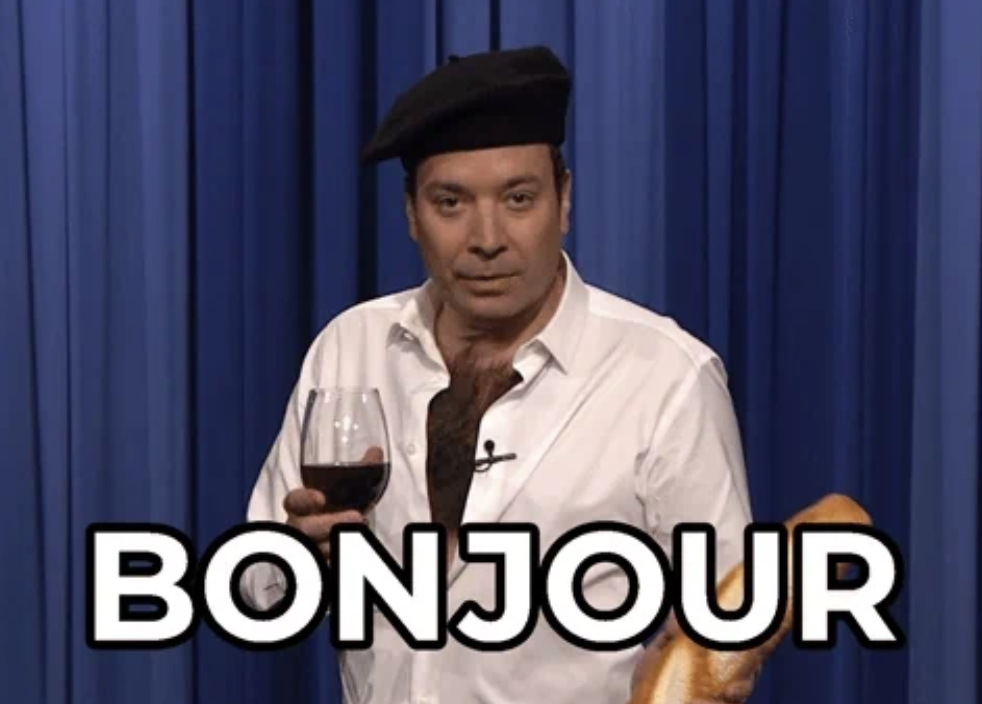 Jimmy Fallon wearing a beret holds a baguette and a glass of wine on a stage. The word "BONJOUR" appears in large text at the bottom