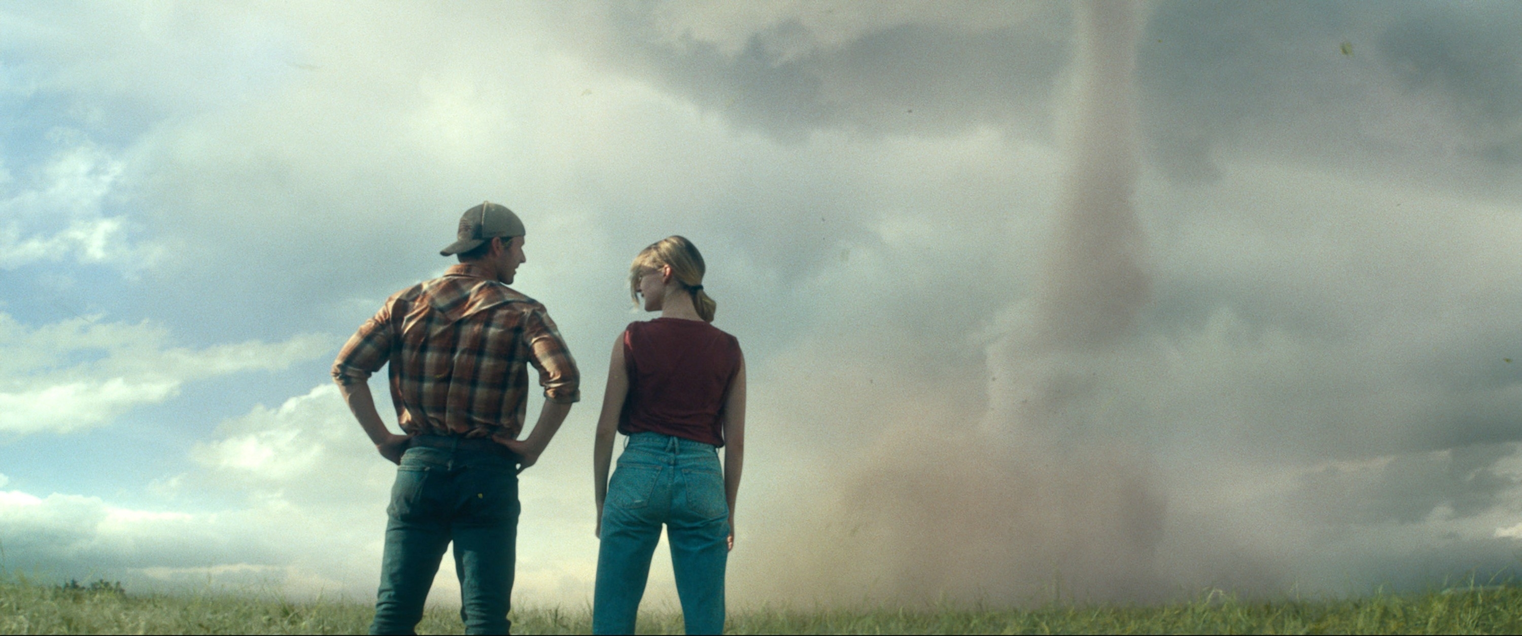 Two people in casual clothing stand in a field, watching a tornado in the distance