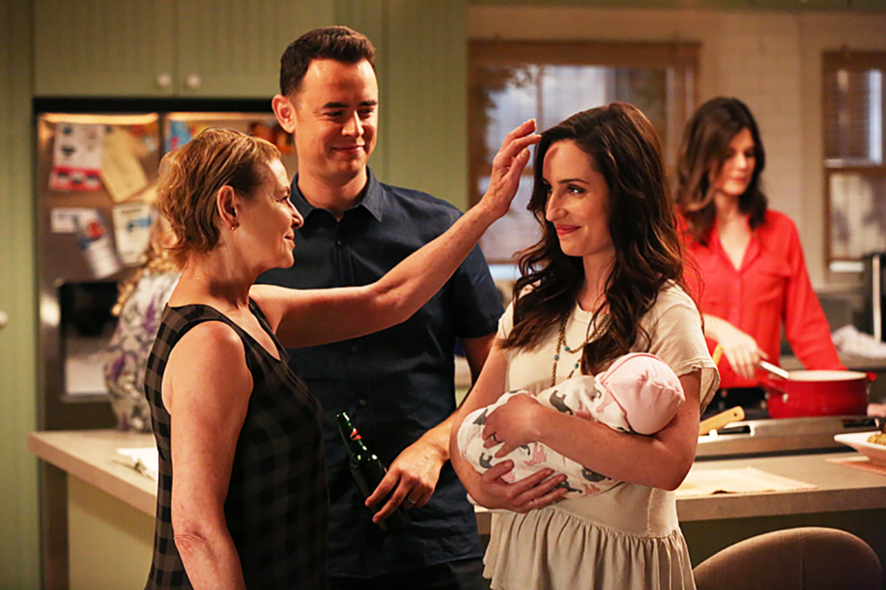 Scene from a TV show with three adults and one newborn. A woman in a checked top smiles and touches another woman's head while a man in a polo shirt looks on