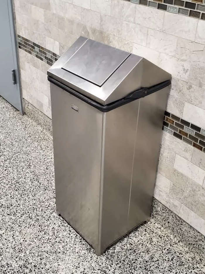 A tall, stainless steel trash can with a swing lid is placed against a tiled wall in an indoor setting