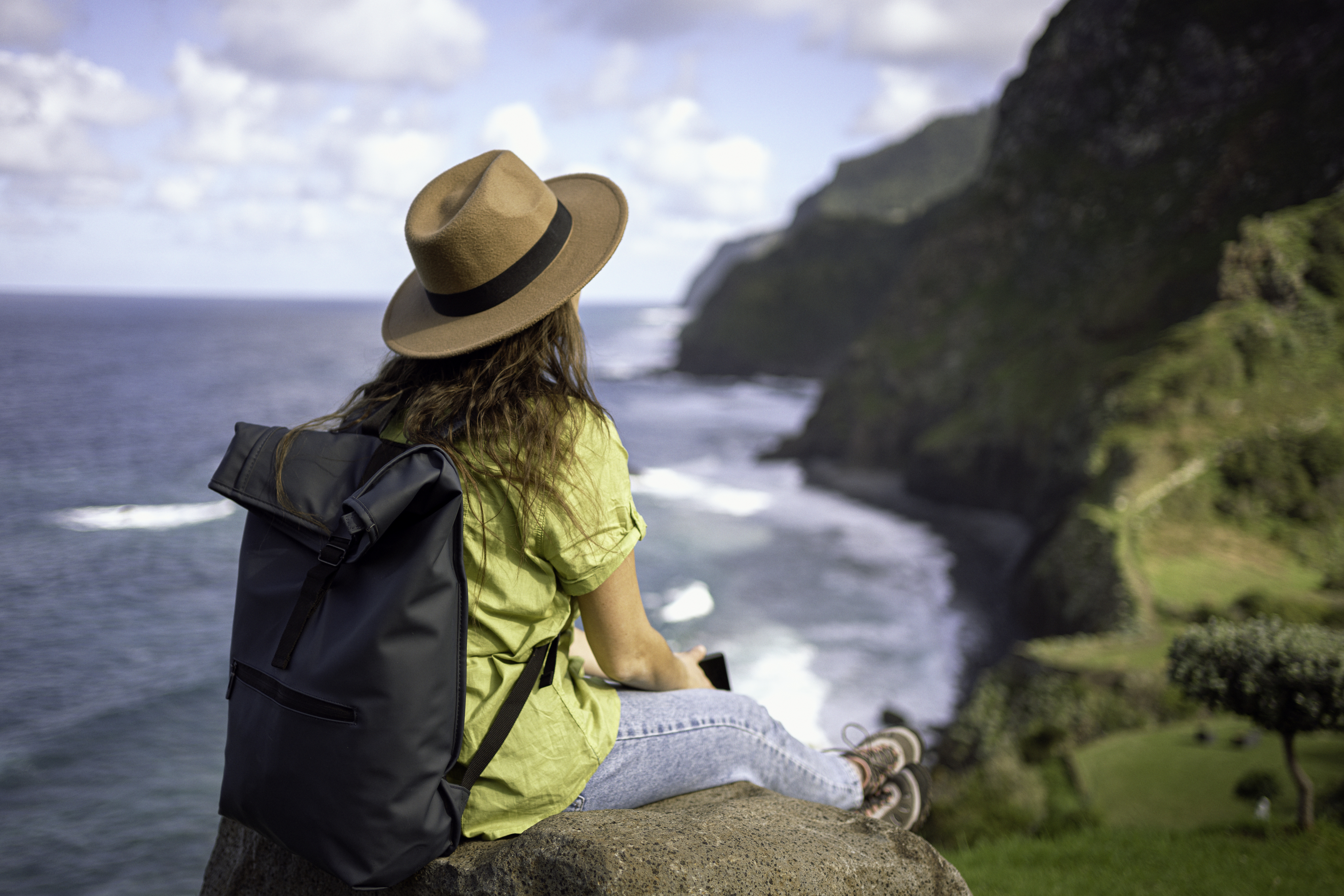 A person wearing a hat and backpack sits on a rock overlooking an ocean with waves and cliffs in the background
