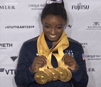Simone Biles stands smiling, holding five gold medals around her neck, in front of a press backdrop. She’s wearing a Team USA jacket
